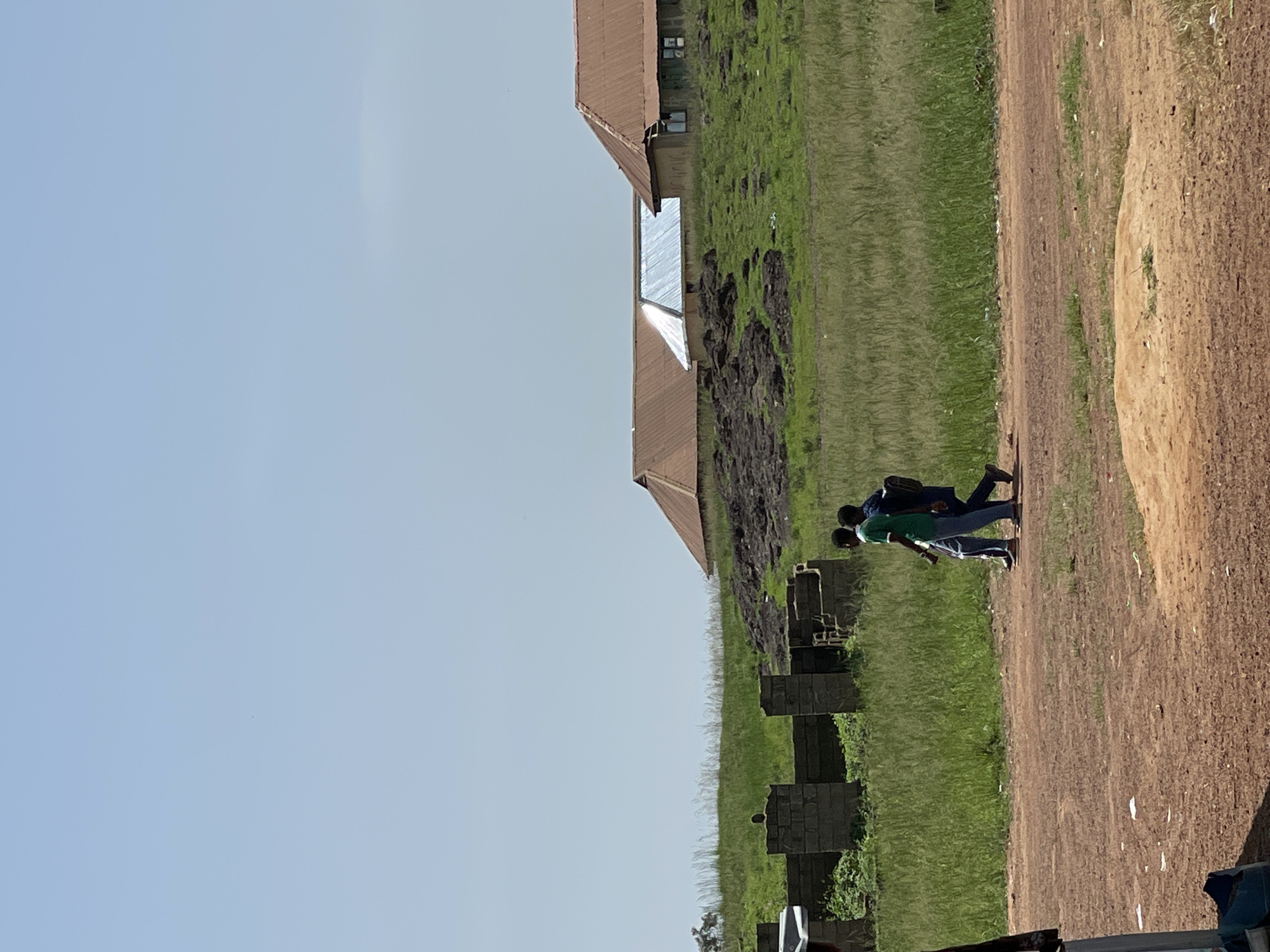 Two young Black males are walking on a dirt path in Durumi, Nigeria. The male on the left is wearing a green t-shirt and blue jeans. The male on the right is wearing a dark blue t-shirt and dark pants, and he is carrying a backpack. They are walking towards the right side of the image, away from the viewer. The environment is dry and dusty, with sparse vegetation. In the background, there are some unfinished brick structures and several buildings with corrugated metal roofs, some of which appear to be residential or part of a small settlement. The sky is clear and light blue, indicating a sunny day. The scene appears to be in a rural or semi-rural area, possibly on the outskirts of a town or village.