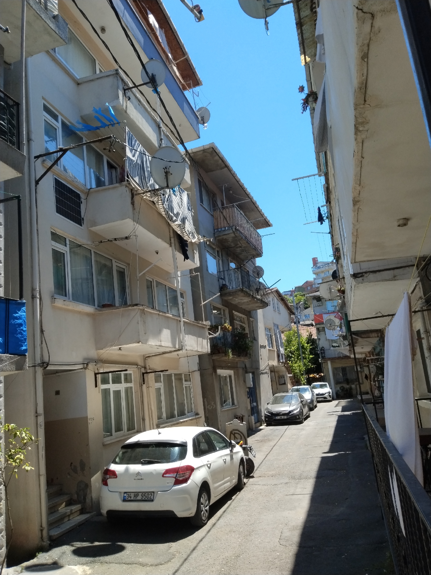 The image depicts a narrow street in an urban area, likely in an unnamed city of unknown location. The street is lined with multi-story buildings of similar architectural style, predominantly light-colored with balconies and windows. Several satellite dishes are mounted on the buildings' facades. A white car is parked on the left side of the street, and two more cars can be seen further down the street. Laundry lines are strung between buildings, and various items are hanging from the balconies. The sky is clear and blue. The lighting suggests it is daytime.