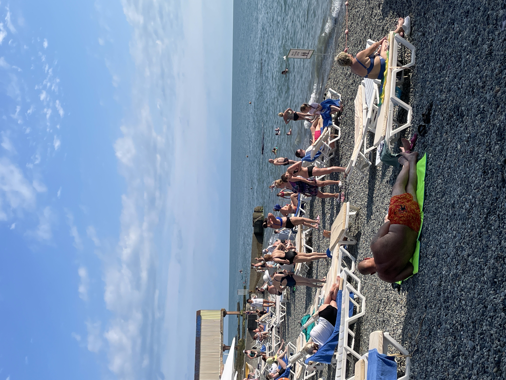 This is an eye-level, wide shot of a crowded beach in Sochi, Russia. The scene captures people sunbathing on lounge chairs and towels on a pebble beach, with the calm, blue sea stretching to the horizon under a partly cloudy sky. Several individuals are seen swimming or wading in the water. A building with a striped facade stands on a raised concrete structure to the left, and a sign with pictograms is visible near the water's edge on the right. The overall atmosphere is one of leisure and vacation.