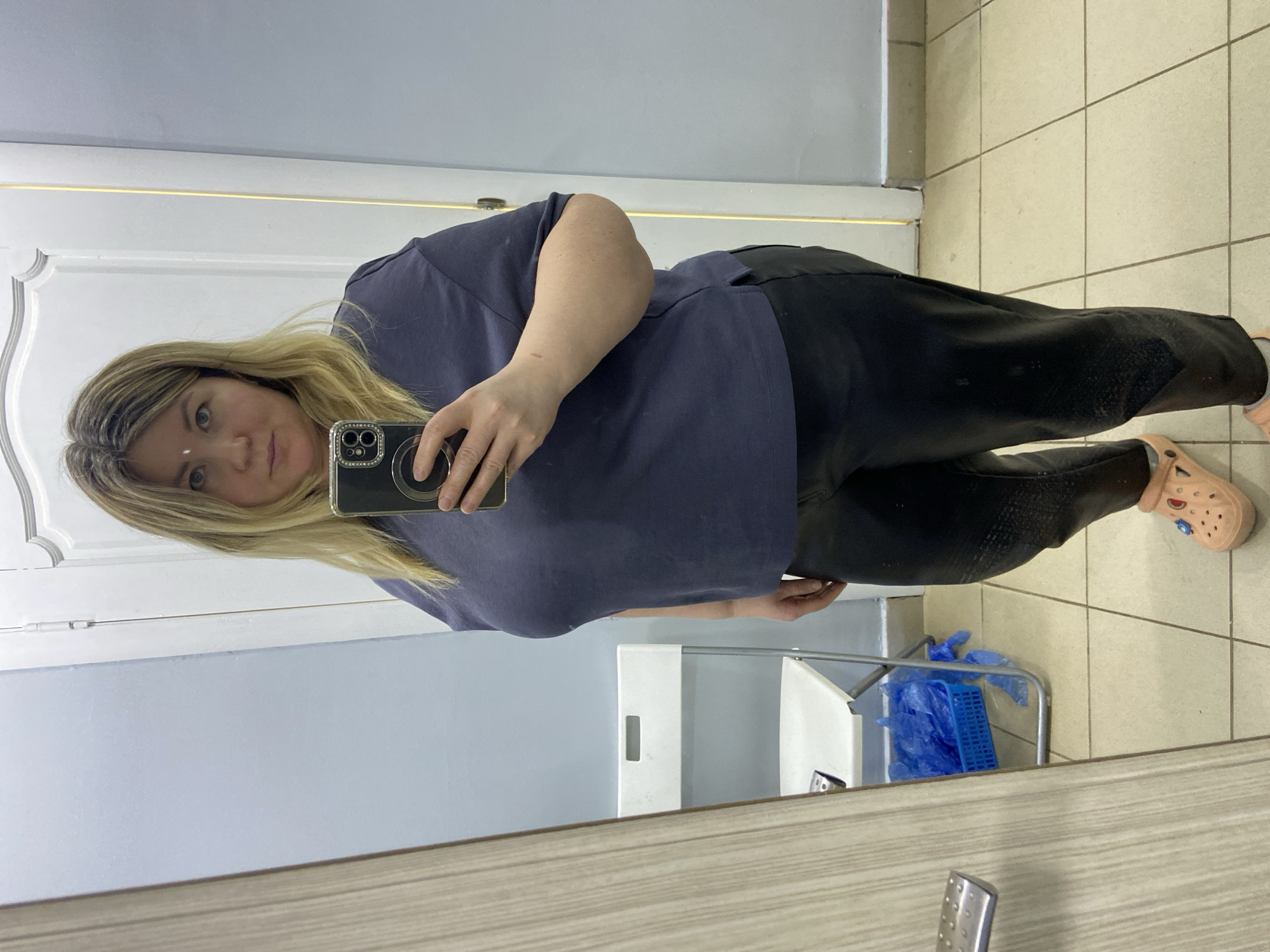 A blonde woman is captured in a mirror selfie, holding a smartphone with a clear case in front of her. She wears a dark blue, loose-fitting top and black pants, paired with light peach Crocs that have decorative pins on them. A small white dot is visible on her forehead, between her eyebrows.
The indoor setting features a white door with decorative molding on the left side of the reflection, and a light blue wall above it. The floor is tiled with light-colored square tiles and darker grout lines. In the lower right of the reflection, a white chair or stool is partially visible, with a blue plastic basket filled with crumpled blue plastic bags beneath it. The scene suggests a casual, functional space, possibly a changing room or a public area. No discernible text is visible in the image.