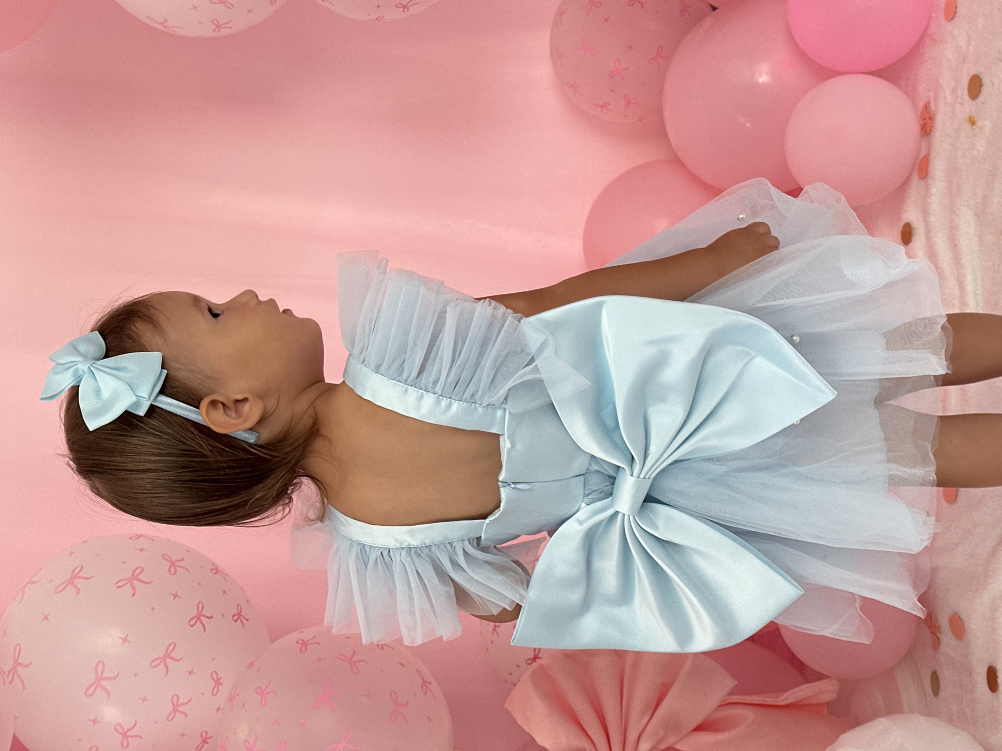 In this vertical, medium shot, a baby is dressed in a light blue, satin and tulle dress with a large satin bow on the front. A matching blue bow is also on her head. She is lying down on a pink background adorned with balloons. Some balloons are solid pink, while others are light pink with a repeating pattern of pink bows and stars. The overall setting suggests a celebration or birthday party. The lighting is soft and even, indicating an indoor setting with professional lighting. There is no visible text in the image. The baby's face is turned to the side, and her expression is neutral. The focus is on the baby and her festive attire. The scene evokes a sense of innocence and celebration.