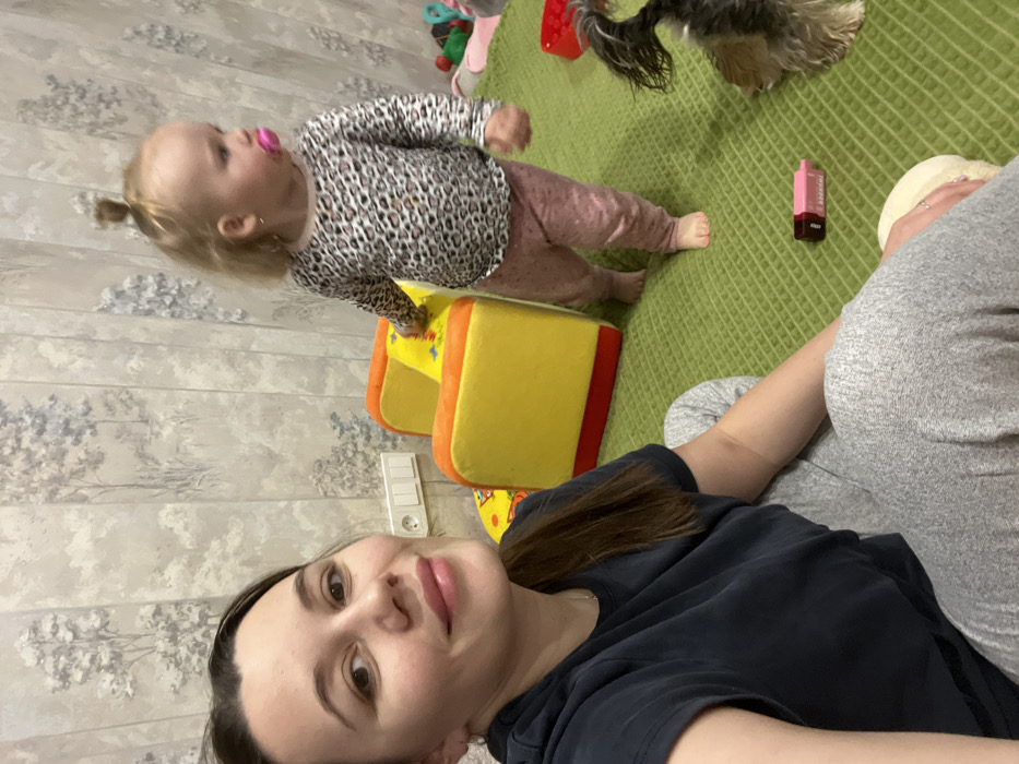 An indoor domestic scene features three subjects: an adult female, a toddler, and a small dog. The adult female, with long brown hair, occupies the left foreground, dressed in a dark short-sleeved top and light grey sweatpants, holding a device to capture a selfie as she looks directly at the camera with a slight smile. A fair-haired toddler, wearing a leopard print long-sleeved shirt, pink pants, and a pink pacifier, stands barefoot in the midground on the right, looking away from the camera. Her left hand rests on a small, yellow and orange children's armchair. On the far right, partially visible, is a small dog, possibly a Yorkshire Terrier or similar breed, facing downwards. A pink and black vape pen lies on the green, textured rug between the adult and the dog. The background wall is covered with light-toned wallpaper featuring a grey and white tree pattern, with an electrical outlet and two switches visible. Other colorful children's toys are present on the rug in the background. The location is an unspecified indoor residence; no information within the image details a specific location in Leninakan, Russia.