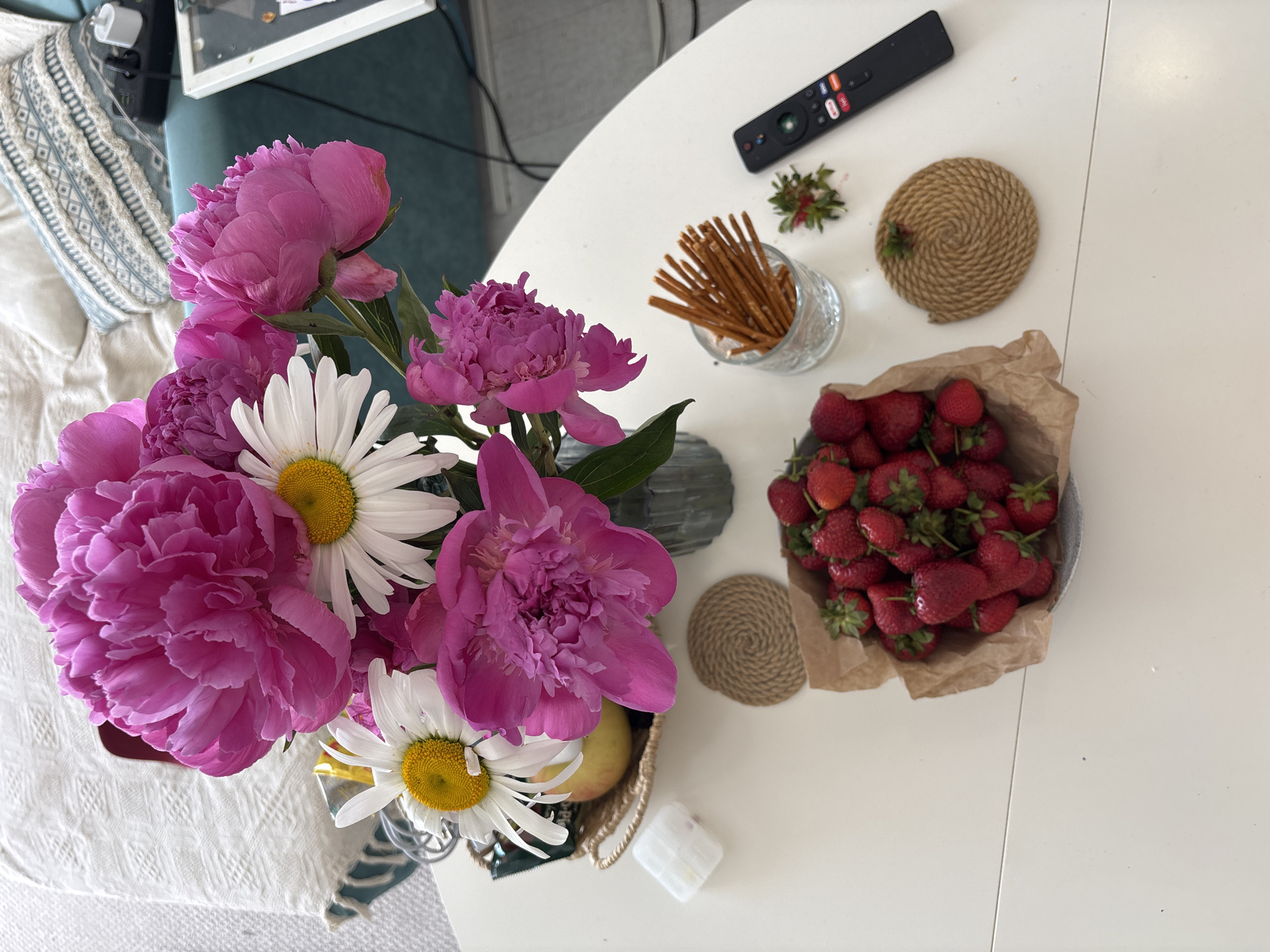 A high-angle shot shows a white table with a bouquet of pink peonies and white daisies in a vase. Next to the vase are pretzel sticks in a glass, two woven coasters, and a metal bowl filled with strawberries on a piece of brown paper. A black remote control is also visible on the table. Part of a pillow with a blue patterned fabric and a teal seat are visible. The location and city cannot be determined from the image.