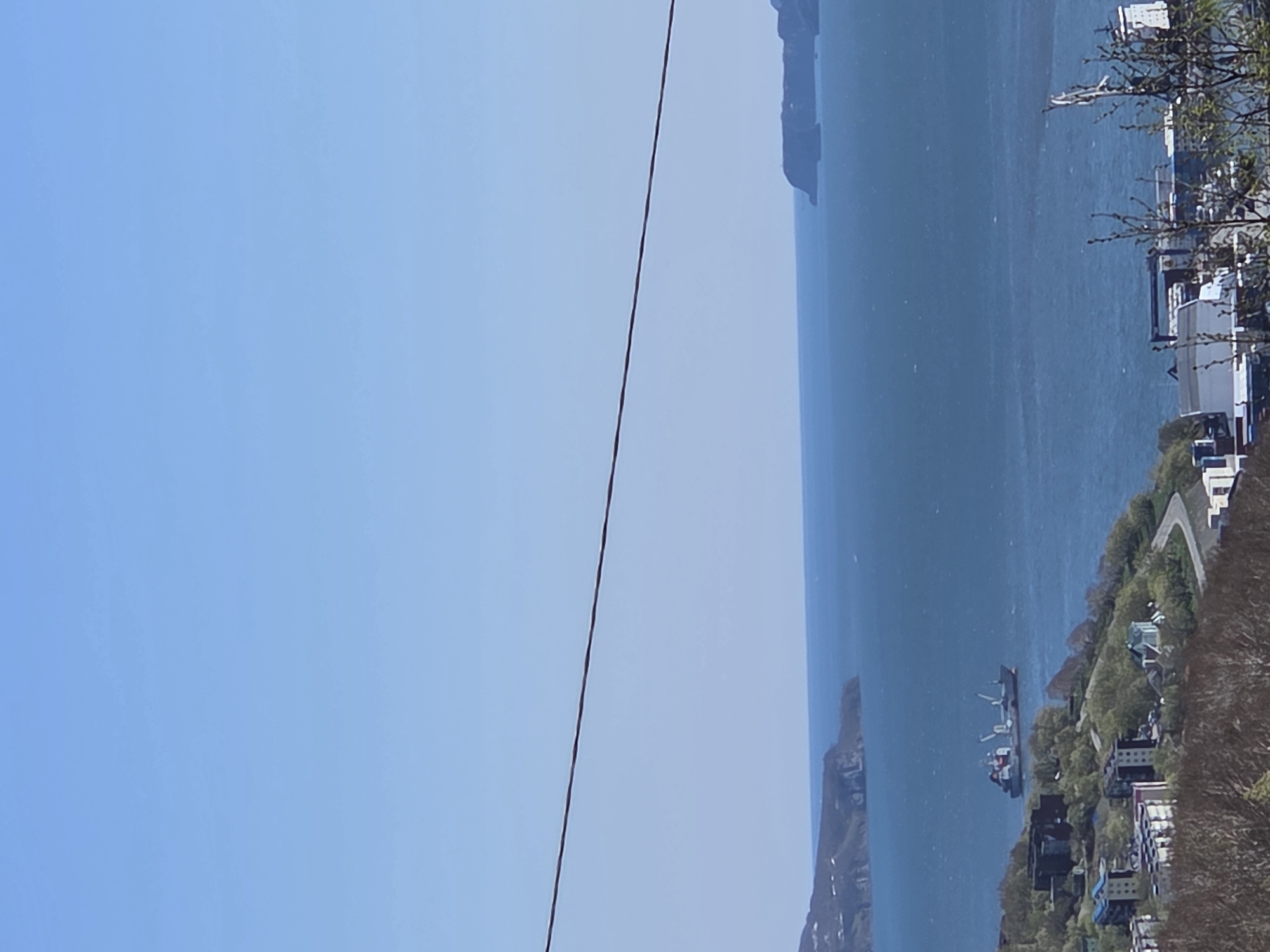 The image depicts a coastal scene in Unknown. A body of water, possibly the ocean, dominates the right side of the image, with landmasses visible in the distance and along the shoreline. Buildings and trees are situated along the coast. A power line runs diagonally across the sky, from the upper right corner to the lower left corner. The sky is a clear blue. A cargo ship is in the water. The photograph is slightly tilted counter-clockwise.