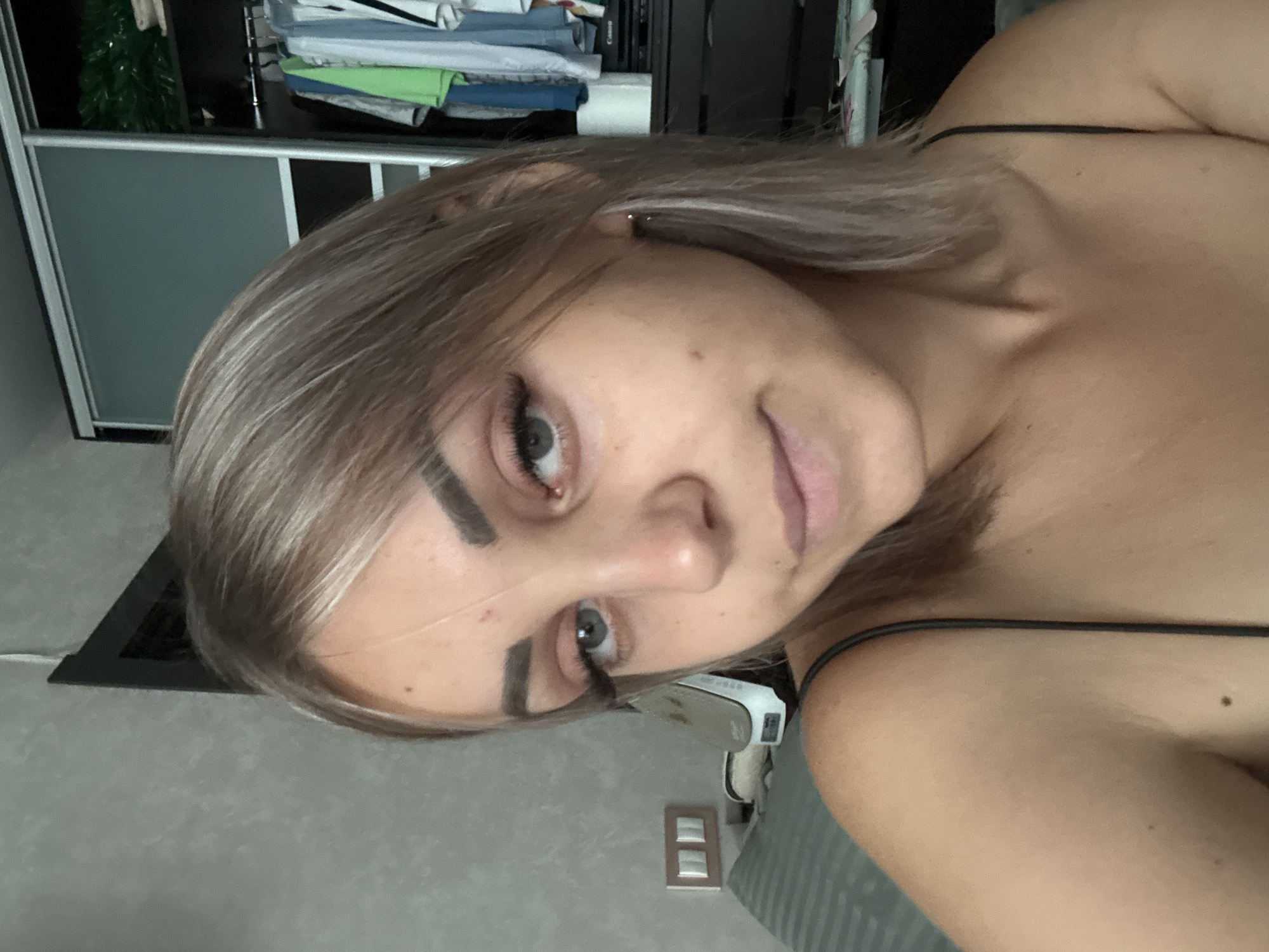 A young woman with blonde hair is captured in a close-up, selfie-style shot. She is positioned in a room with a gray, textured wall as the backdrop. To her right, a large sliding door wardrobe is visible, containing an assortment of clothing on hangers. In the background, a dresser with several drawers is present, and to the left, a power outlet with two switches is mounted on the wall. The woman is wearing a dark-colored spaghetti strap top. The lighting suggests an indoor setting, possibly a bedroom. The image does not provide enough information to determine the specific location within Yuzhno-Sakhalinsk, Russia.