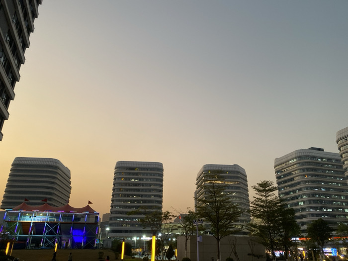 The image depicts an urban scene in Guang Zhou Shi, China, captured during dusk or twilight hours. The sky transitions from a warm, golden-orange near the horizon to a lighter blue-grey overhead, indicating the time of day.
The scene is dominated by several modern, high-rise buildings featuring distinctive rounded corners and horizontal window bands. One building is partially visible on the far left, while four additional similar structures are prominent in the mid-ground and background, with internal lights illuminated.
In the lower-left quadrant, a large, multi-level temporary structure stands out. It has a tent-like roof with sections of red and purple fabric and is brightly illuminated by vibrant blue and purple lights, suggesting an ongoing event, fair, or festival. Scattered trees and other low vegetation are visible in the foreground and between the buildings.
On the ground level, several indistinct figures of people are present, some near the colorful temporary structure and others further to the right, implying a public gathering or evening stroll. Yellow ground-level pole lights or fixtures also contribute to the artificial illumination of the area. No discernible text is visible on any signs or structures within the image. The overall atmosphere suggests a lively evening in a modern city.