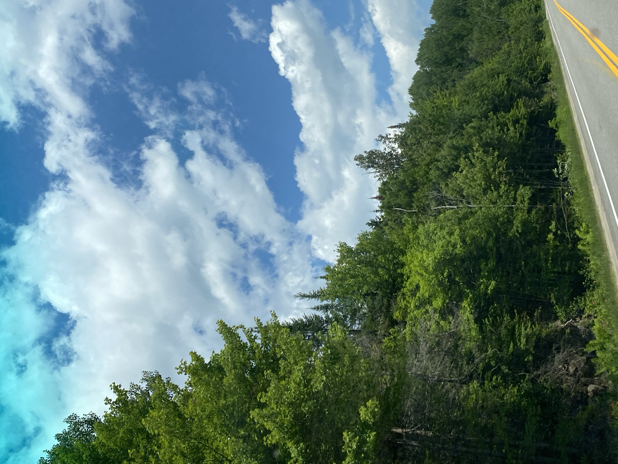 The image shows a paved road with yellow double lines, curving to the right, bordered by a dense forest of green trees on the left. The sky above is blue with scattered white cumulus clouds, and a turquoise hue in the upper left corner. The trees are lush and appear to be in full summer foliage. The terrain slopes upwards from the road into the forest, with rocky outcrops visible in places. The scene appears to be a natural, rural setting, likely a highway or scenic route. There are no people or structures visible in the image, suggesting a remote or undeveloped area. The image was taken in the city of Lac-Simon, Canada.