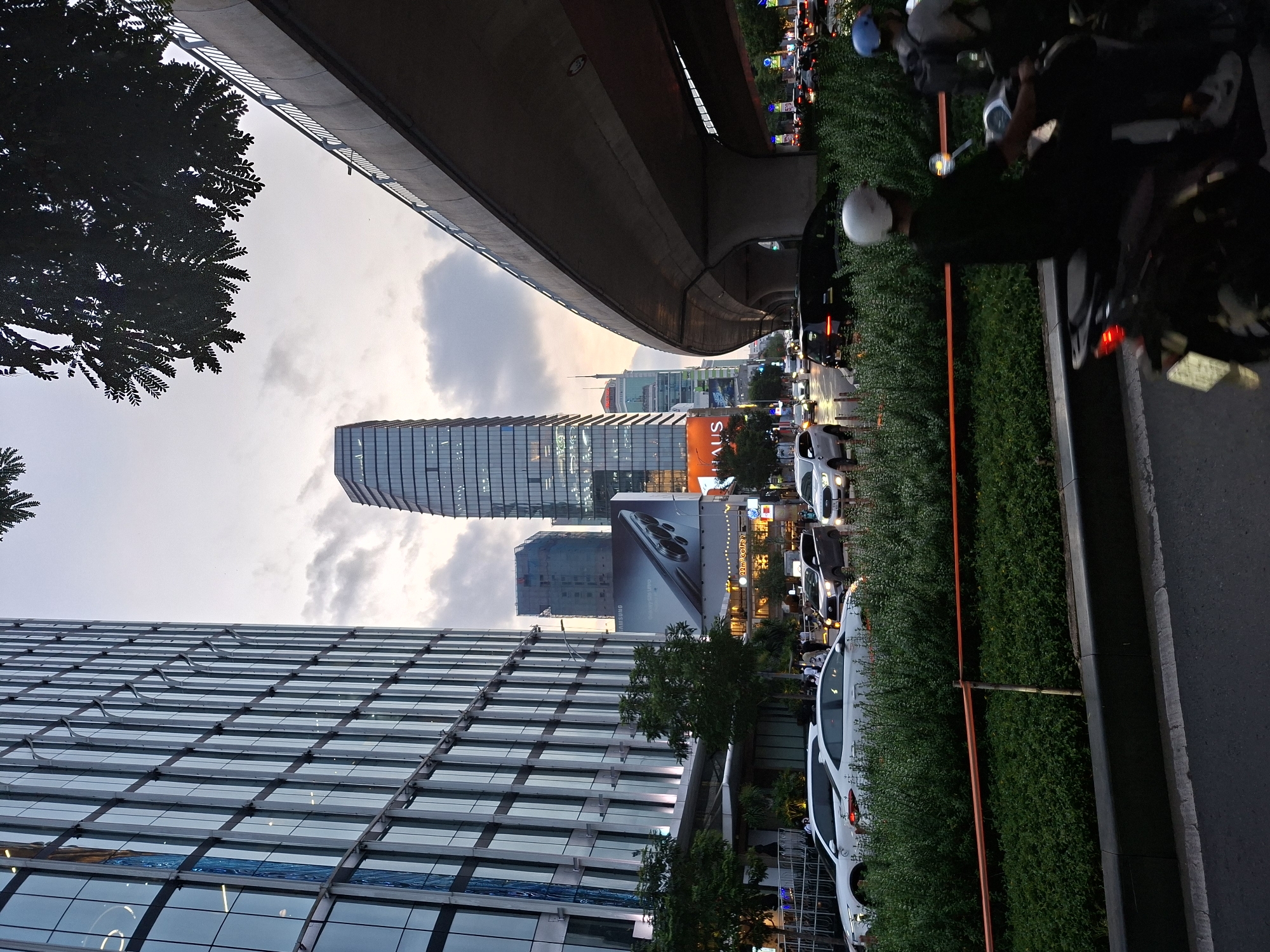 This image depicts an urban scene in Thủ Đức, Vietnam, likely during the evening or dusk, as indicated by the artificial lighting and the soft, diffused natural light. The perspective is unusual, with the camera tilted at an angle, making it appear as if the viewer is looking up at the cityscape.
The foreground is dominated by a modern glass-fronted building, its facade reflecting the surrounding environment and creating a grid-like pattern. To the right of this building, a street runs parallel to a lush green hedge. Traffic is visible on the street, with cars and motorcycles forming a queue, suggesting moderate congestion. Several people are on motorcycles, some wearing helmets.
In the background, a distinctive skyscraper with a curved, segmented facade stands tall, illuminated by internal lights. Below it, a large billboard displays a smartphone, possibly an advertisement for Samsung. The overall setting is a busy urban environment with a mix of contemporary architecture and infrastructure, including what appears to be an elevated road or bridge structure arching overhead. The sky is cloudy, with hues of orange and grey suggesting the transition from day to night. The scene is alive with activity, characteristic of a bustling city. Visible text includes "Samsung" and "HAUS".