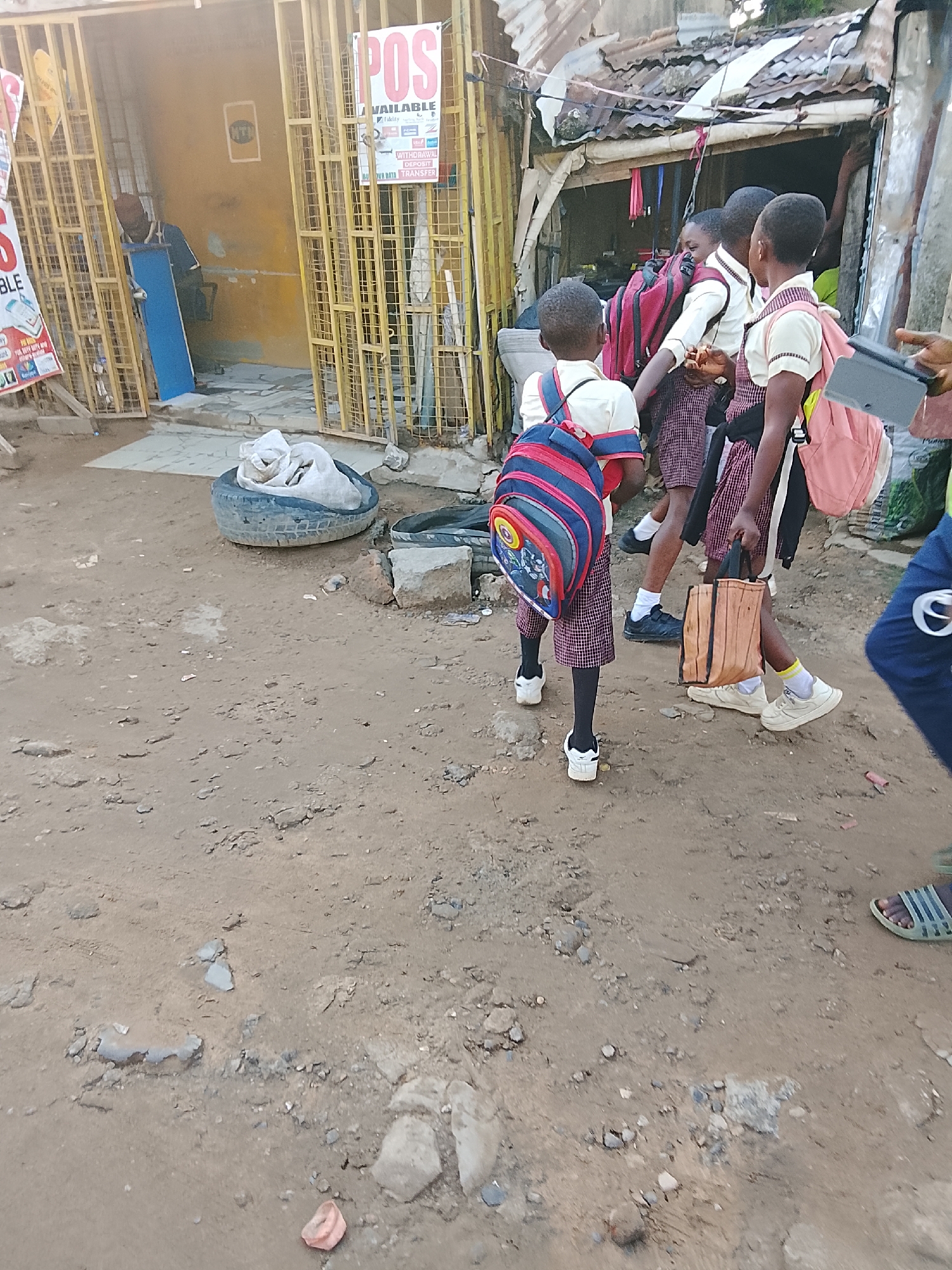 This image captures an outdoor scene featuring several young children, likely students, in what appears to be a bustling, semi-urban area, consistent with Abuja, Nigeria. The ground is unpaved, consisting of reddish-brown dirt and scattered rocks and debris.
The main subjects are approximately four to five children, dressed in school uniforms of white shirts and plaid (red and white) skirts or shorts. They are all carrying school bags: one child in the foreground has a distinctive red and blue striped backpack, another wears a red backpack, and a third carries a light pink one. One child also holds a light brown bag with a wood-grain pattern and appears to be holding a small tablet or electronic device. They are walking together, seemingly interacting or conversing, possibly on their way to or from school. Their footwear includes white sneakers, black shoes, and white socks. Another person, partially visible on the far right, wears blue pants and a striped slide.
In the background, there are informal structures and stalls. On the left, a yellow-painted shop or stall is secured by yellow metal gates. Inside, a person wearing a dark shirt and a red cap is partially visible, holding a blue panel. A prominent sign on this stall advertises "POS AVAILABLE," listing services such as "WITHDRAWAL," "DEPOSIT," "BUY DATA," and "TRANSFER," along with logos of various Nigerian banks and payment services like Fidelity, GTBank, Zenith Bank, UBA, Access Bank, First Bank, and Opay. To the right, another building with a weathered corrugated metal roof and a dark opening suggests a dwelling or another shop.
On the ground in the mid-ground, discarded tires are visible, one containing a white sack. The lighting appears soft, suggesting either early morning or late afternoon. The overall scene depicts a glimpse into daily life in a local community.