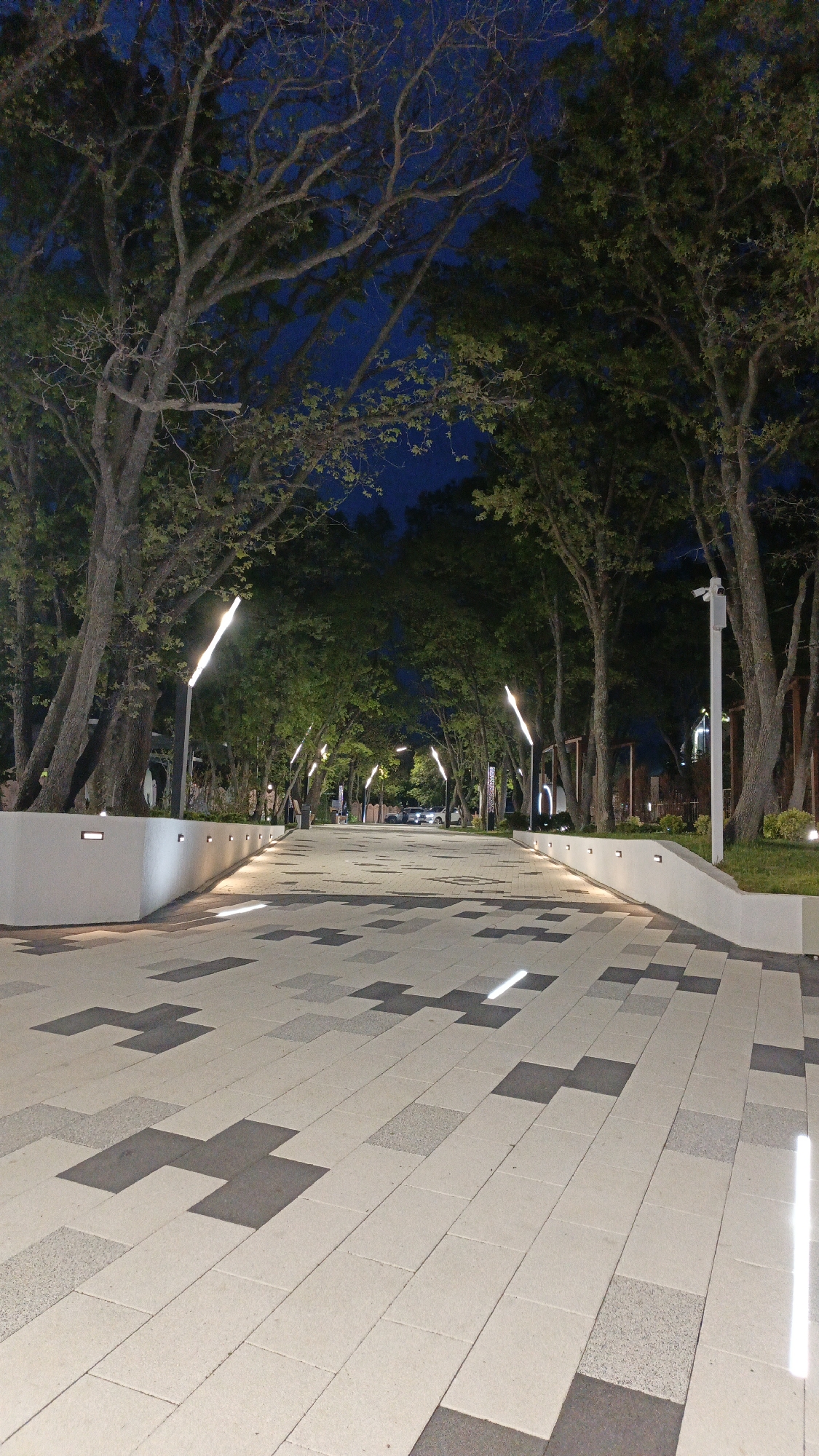 The image depicts a well-lit pedestrian walkway in Kabardinka, Russia at night. The walkway is paved with a pattern of light and dark rectangular stones, some arranged in cross shapes. Low concrete walls border the walkway on either side, with embedded lights illuminating the path. Trees line the walkway, their branches reaching overhead, creating a canopy. Tall, slender light poles are spaced along the path, casting a bright glow. Further down the path, vehicles are visible. A security camera is mounted on a pole on the right side of the walkway. The sky is a deep blue, suggesting it is late evening.