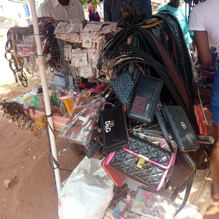This image depicts an outdoor vendor stall situated in Nsukka, Nigeria. The stall showcases a variety of merchandise. On the left side, numerous necklaces, bracelets, and earrings are displayed, many individually sealed in clear plastic packaging. Also present on this side are bundles of colorful hair ties or scrunchies. The right side of the stall features an assortment of handbags, wallets, and belts, with several items exhibiting visible brand logos, including "D&G." The goods are arranged on a simple stand structure, supported by white poles.
In the immediate background, the upper torso of an adult male wearing a light-colored shirt is visible, positioned behind the stall. To the far right, the lower torso and bare foot of a person in blue shorts are captured, indicating an individual standing adjacent to the display. The ground surface is reddish-brown earth. In the distant background, several indistinct figures and structures, including a signpost, are visible, suggesting a street or public market area.