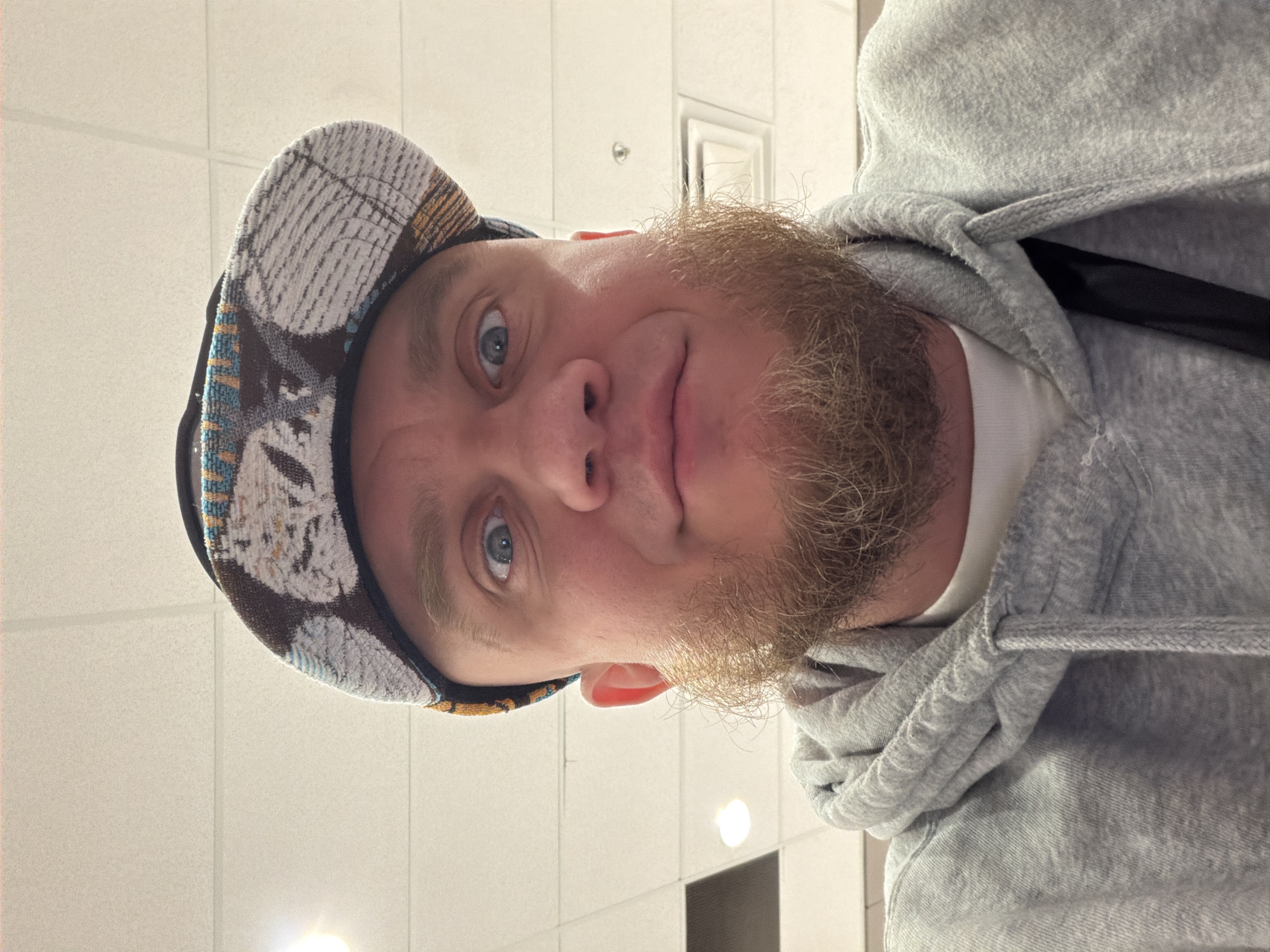A selfie captures a man with blue eyes, a light complexion, and a full beard. He wears a patterned hat with a black brim and a gray hooded sweatshirt. The background is a white tiled ceiling with a rectangular light fixture. The photograph appears to be taken indoors. It is not possible to identify the city or location based only on these details.
