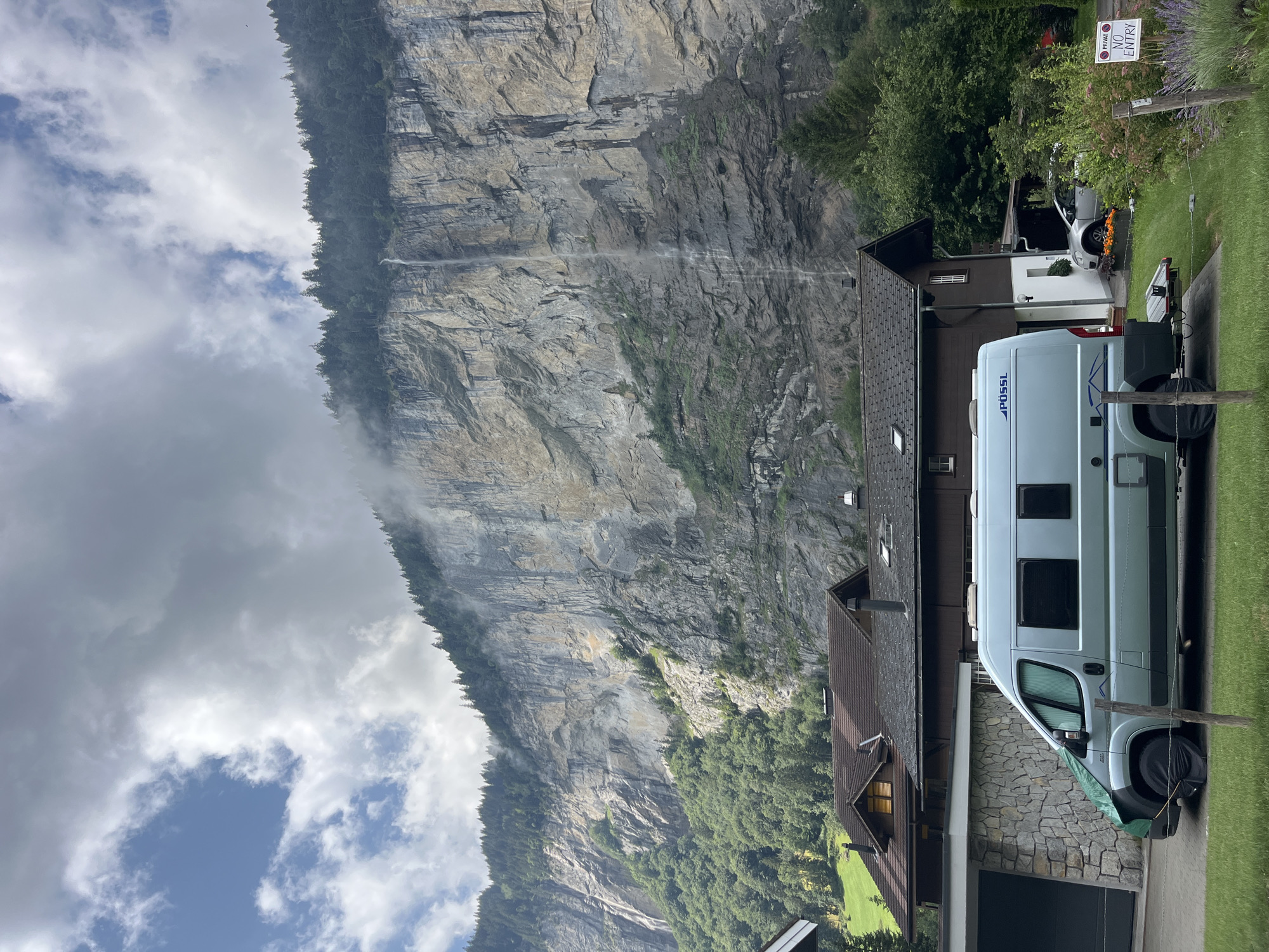 The image depicts a scenic view in Wengen, Switzerland, with a large cliff face dominating the background. A waterfall cascades down the sheer rock, partially obscured by mist and clouds. Dense green forest covers the lower slopes of the cliff, transitioning into the rocky expanse above. In the foreground, a light blue camper van is parked on a gravel driveway. Adjacent to the van are two residential buildings with dark brown tiled roofs. A stone-clad garage is to the left of the van, and a silver car is visible in front of one of the houses. A small sign with "PRIVAT NO ENTRY" is posted on a wooden fence in the lower right corner. The sky is a mix of blue and grey clouds.