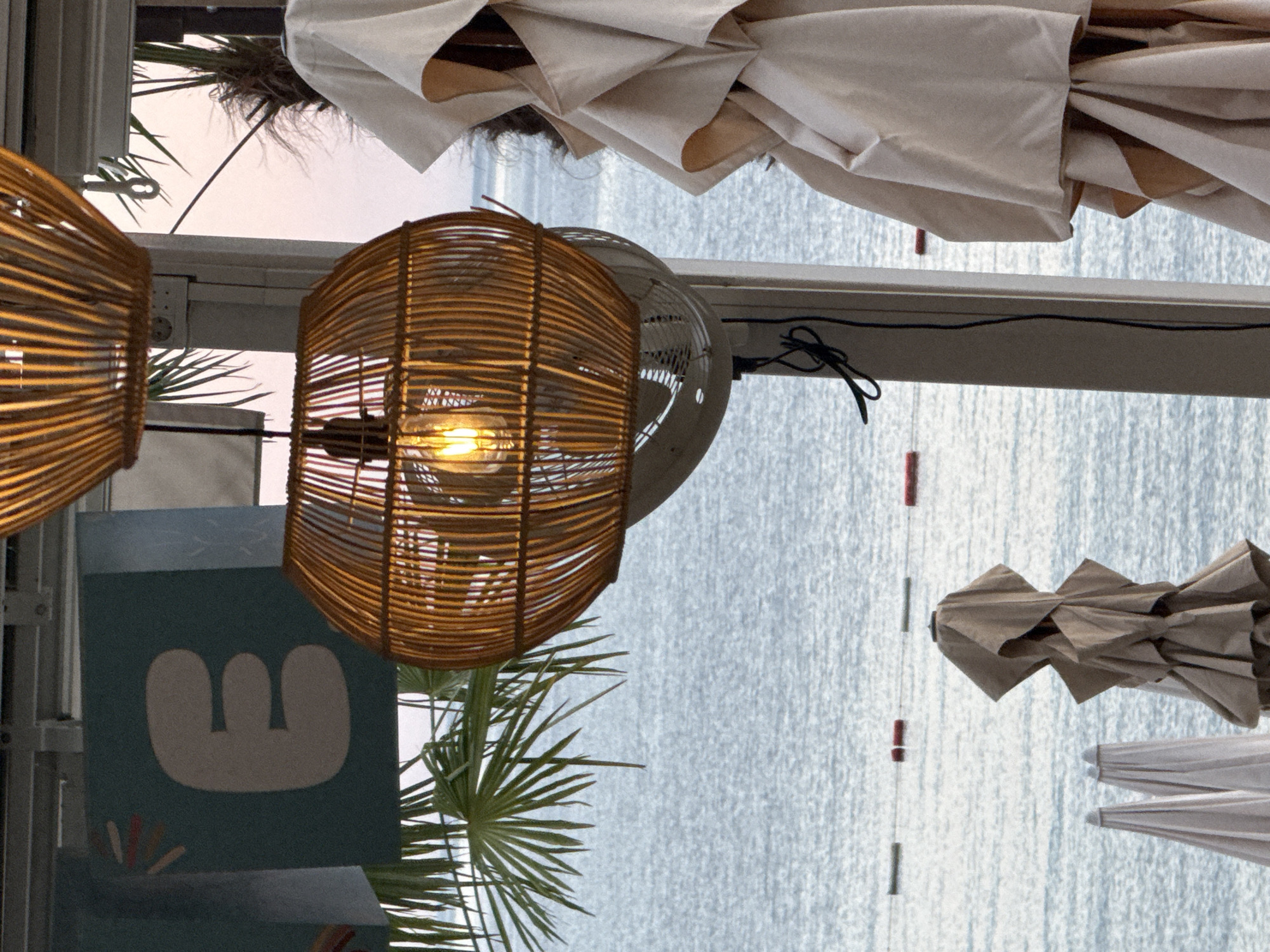 The image depicts a seaside scene in Kamenovo, Montenegro, likely at a beach cafe or resort. In the foreground, several closed beach umbrellas are stacked, with one prominently featured in beige fabric on the right side. Above them, a woven rattan lamp with a lit bulb hangs, casting a warm glow. Behind the umbrellas and lamp, the calm, blue sea stretches towards the horizon. A line of red and green buoys marks a boundary in the water. Palm fronds are visible in the upper left, and a partial view of a sign with a stylized letter "E" on a teal background is also present. The sky above is a soft pink and orange, indicating either sunrise or sunset. A white ceiling fan is partially visible beneath the rattan lamp.