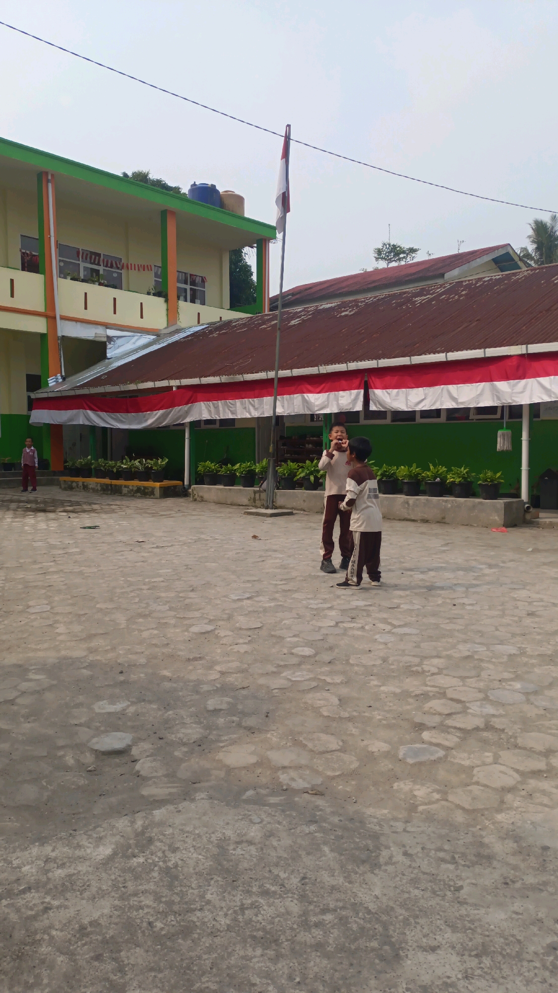 The image shows an outdoor courtyard with two young boys in the foreground, standing on a paved surface. One boy, on the left, is wearing a light-colored shirt with brown accents and dark brown pants. He appears to be speaking or shouting with his mouth open. The second boy, on the right, is wearing a similar light-colored shirt and dark brown pants, and is facing the first boy. In the background, a building with a green and orange facade is visible, along with a red and white banner draped across the front. A flagpole with the Indonesian flag is positioned to the right of the building. Several potted plants are lined up along a low wall in front of the building. A third, smaller child in a light-colored shirt and dark pants is visible further to the left, near the building's entrance. The environment appears to be a school or public space. The specific location in Padang Panjang, Indonesia, is not explicitly identifiable from the visual information alone.