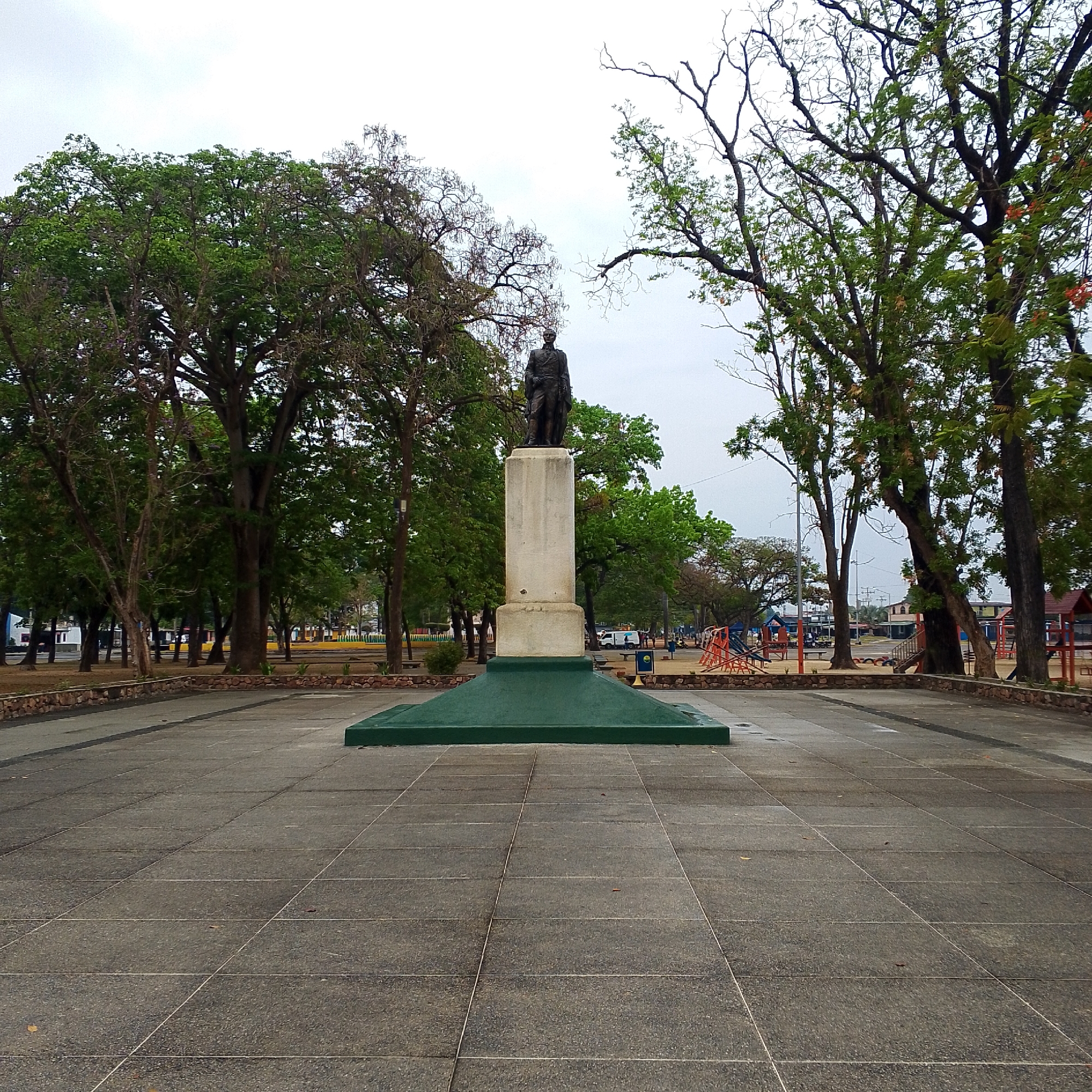 The image captures a static scene of a monument in a public park, likely in San Carlos, Venezuela. In the foreground, a large, dark-colored statue of a man stands prominently on a tall, light-colored rectangular pedestal. The man is depicted in a formal, possibly military, uniform. The pedestal rests on a wide, green, pyramid-shaped base. The ground surrounding the monument is paved with large, grey square slabs.
Numerous large trees with dense green foliage, interspersed with some having sparser leaves or bare branches, frame the central monument and extend into the background, creating a natural canopy. To the right, a colorful children's playground with orange and blue climbing structures and slides is visible among the trees. In the distance, through the trees on both sides, glimpses of buildings and a white utility truck can be seen, indicating an urban or semi-urban setting.
The sky is overcast and uniformly grey, suggesting a cloudy day with soft, diffused lighting and no strong shadows. The absence of people in the frame contributes to a calm and quiet atmosphere. No specific activity or event is taking place. A small, unreadable blue sign is visible near the playground equipment.