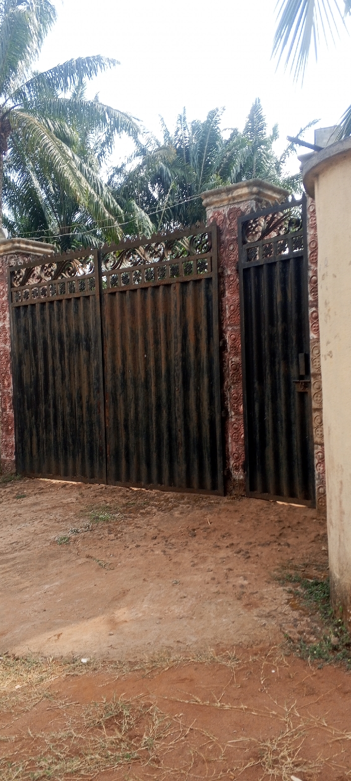 The image displays the exterior of a property entrance in Alor, Nigeria, featuring a metal gate structure. The scene is dominated by a weathered dark corrugated metal double gate, flanked on the right by a narrower, similar pedestrian gate equipped with a latch mechanism. Both gates feature decorative, ornate scrollwork and square patterns along their upper edges. These gates are integrated into reddish-brown pillars and wall sections, textured with a repeating circular or patterned relief design. The tops of the pillars are flat and weathered. To the far right, a light-colored, possibly concrete, cylindrical structure is partially visible. The ground directly in front of the gates consists of reddish-brown dirt or sandy soil, with scattered patches of dry, sparse green and brown vegetation. The background reveals dense green foliage, predominantly palm trees, beneath a bright, overcast sky. No individuals or active subjects are present in the frame.