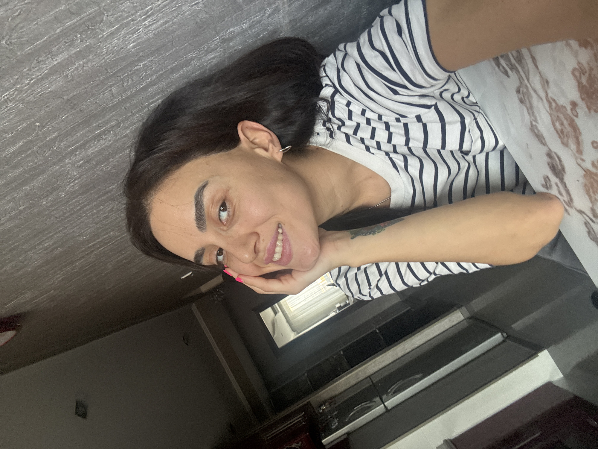 A young woman with dark hair and olive skin, wearing a white and navy striped t-shirt, smiles at the camera. She has her right hand on her cheek, and her left arm rests on a marble-patterned surface. She has bright pink fingernails. A silver hoop earring is visible in her left ear. The background is a textured gray wall. To the left, a dimly lit hallway leads to an open doorway, revealing a bright room with white curtains. A glimpse of a kitchen with stainless steel appliances is visible down the hallway. The overall impression is a casual selfie taken indoors, possibly in an apartment in Tbilisi, Georgia, given the style of the interior.