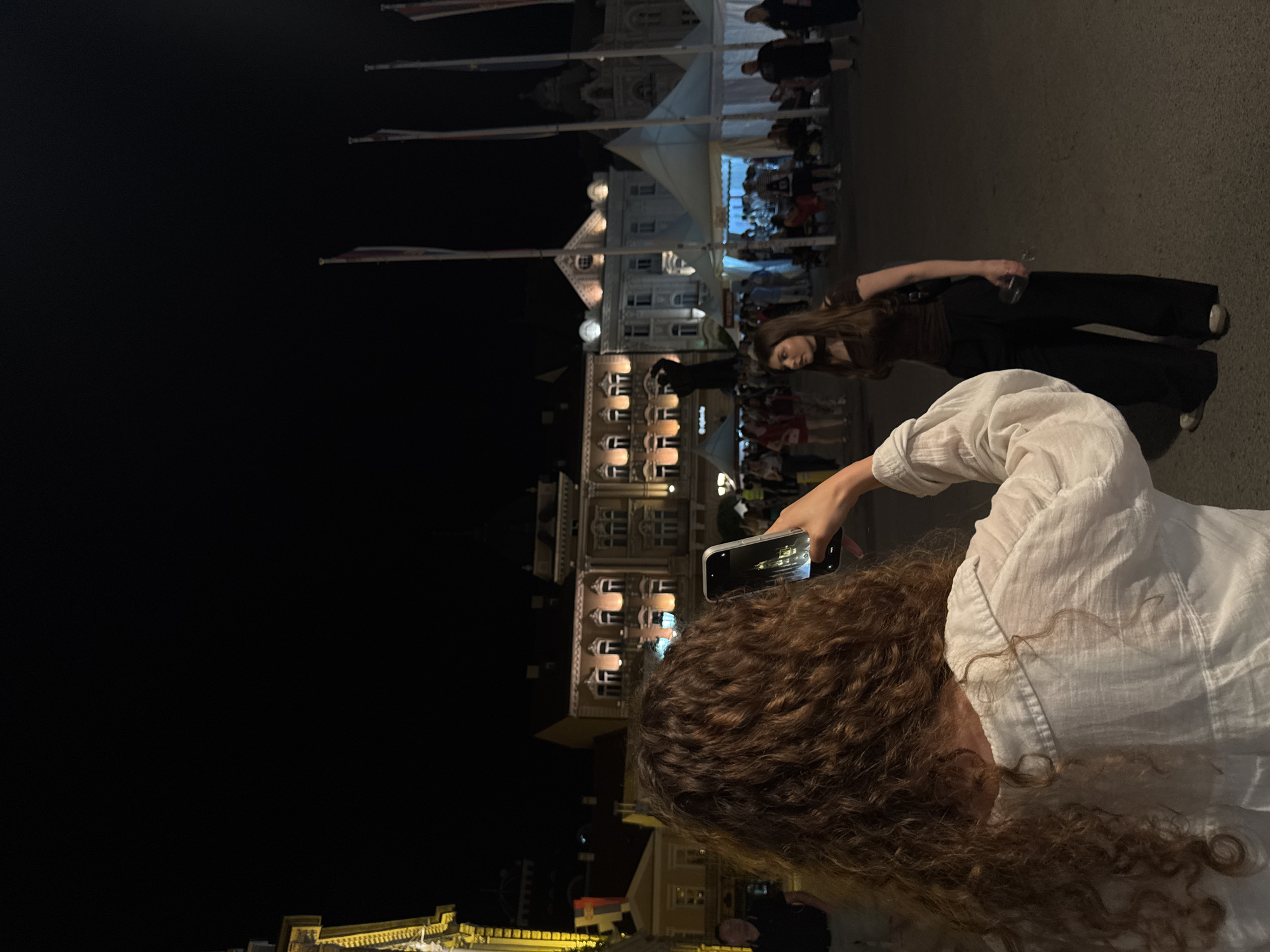 In Novi Sad, Serbia, at night, a person with curly brown hair, wearing a white shirt, holds up a smartphone to take a picture of another person. This second person, a woman with long brown hair, is wearing a dark brown off-the-shoulder top and wide-leg black pants. She is holding a clear wine glass in her right hand. In the background, illuminated buildings with traditional architecture and flags are visible. Several white tents are set up, suggesting an outdoor event or market. People are milling about in the background, and a statue of a person is visible between two buildings. The scene appears to be a public square at night.