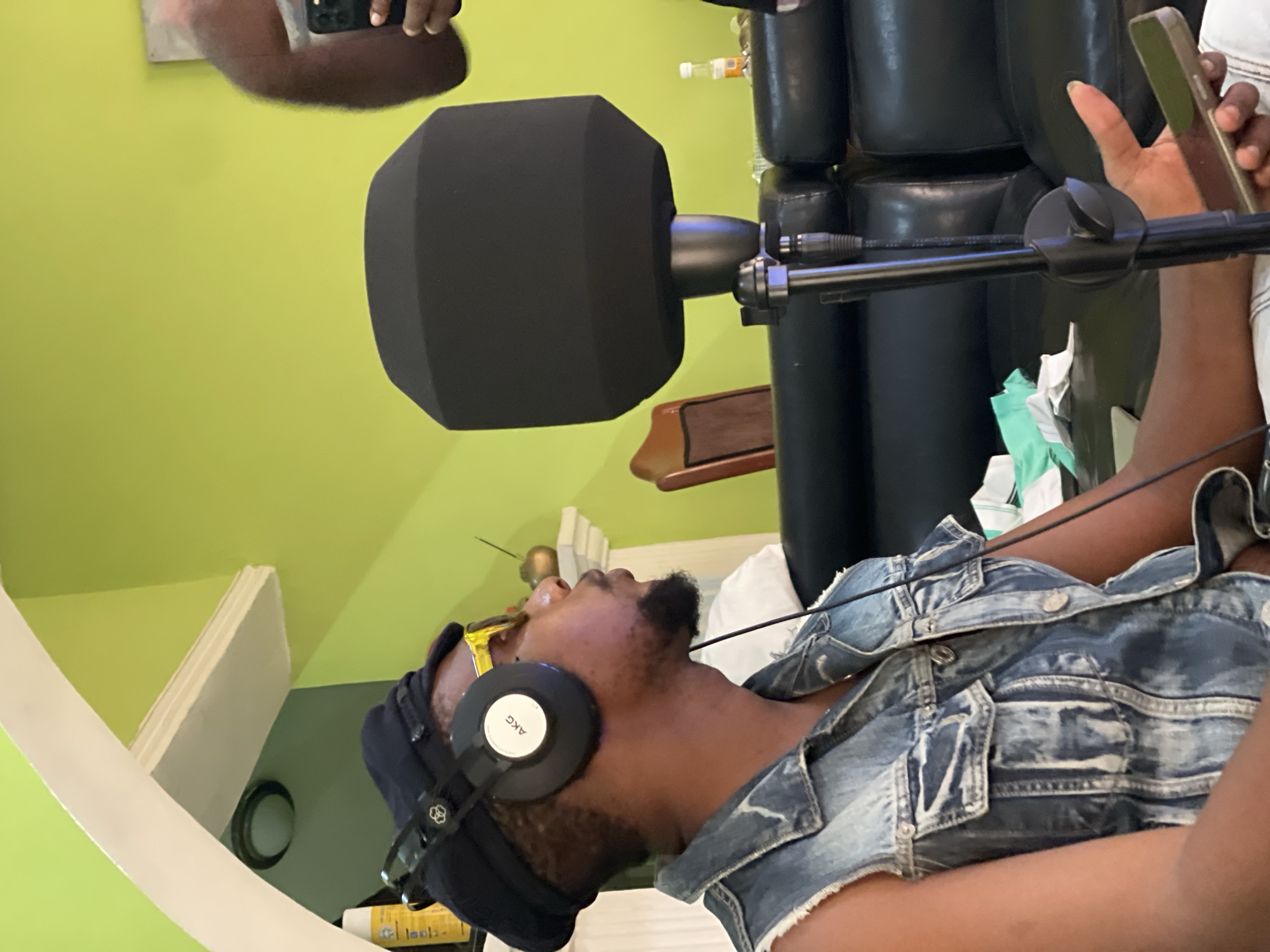 A man with dark skin is wearing AKG headphones, a black beanie, and a denim jacket. He is positioned in front of a microphone with a large, box-shaped foam windscreen, likely for recording audio. The background includes a light green wall, a portion of a black leather chair, and miscellaneous items like a bottle and cables. A hand holding a smartphone is visible, seemingly recording. The location is likely an indoor recording studio. There is no way to confirm the location in Abuja, Nigeria, from the given image.