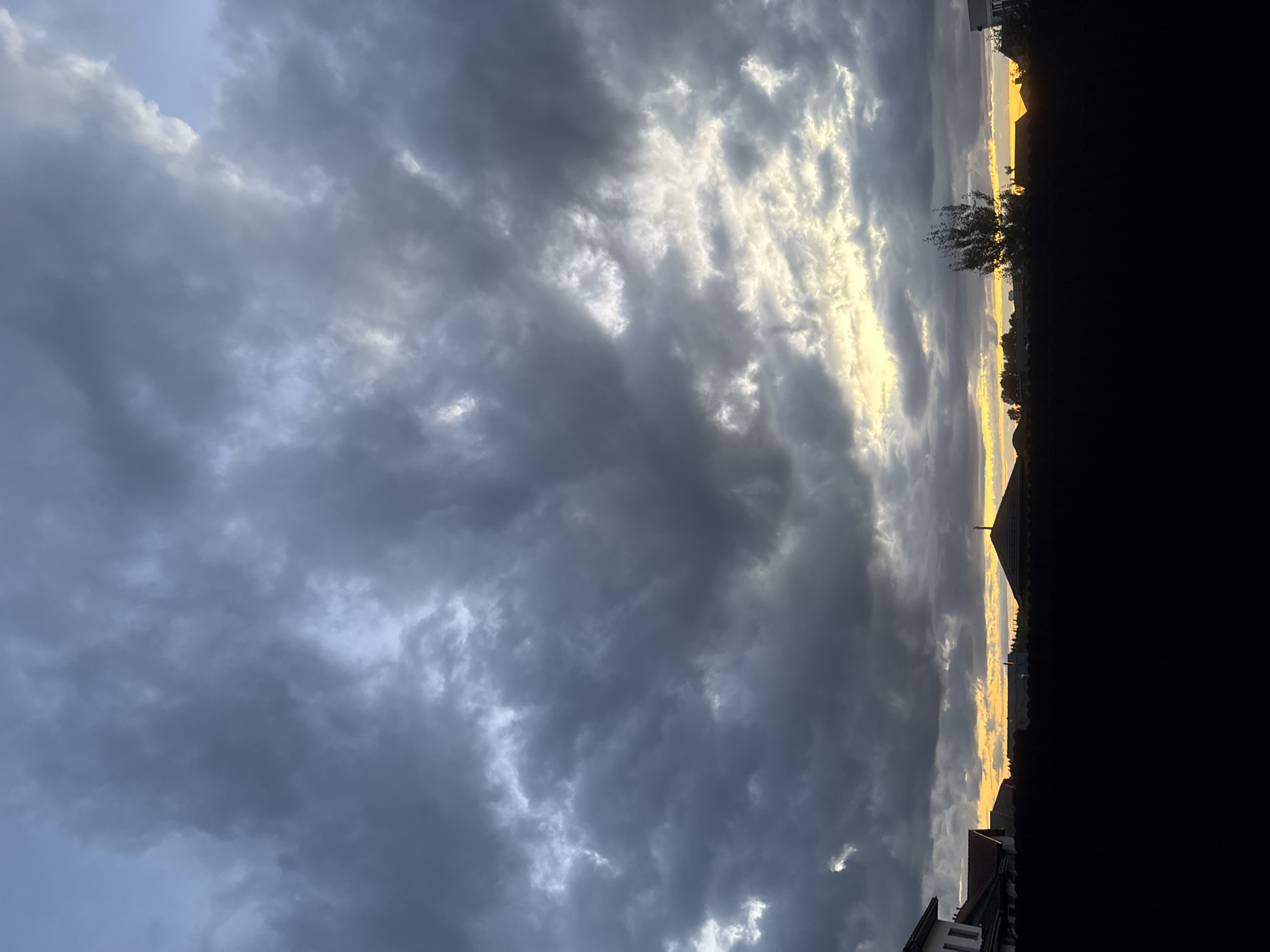 The image captures a dramatic, overcast sky with heavy, dark gray clouds dominating the upper portion. A band of bright yellow and white light peeks through the clouds near the horizon, suggesting either sunrise or sunset. Silhouetted against this luminous sky are the rooftops of houses, a church spire with a cross, and the tops of trees. In the foreground, a solid black silhouette of a fence or hedge runs horizontally across the bottom of the frame. The scene appears to be set in a residential area. Based on the provided location, this is likely a view of houses and a church in the Бараўлянскі сельскі Савет, Belarus, under a moody sky. No people are visible in the image.