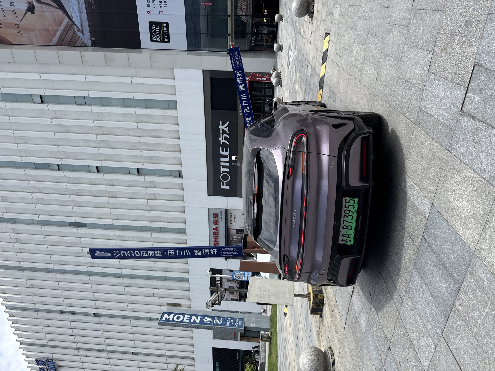 In the Huangpu District of China, a purple Xiaomi SU7 electric vehicle is parked on a paved plaza in front of a modern building with a vertical facade. The car has a green license plate that reads "A B73955". Signs for "MOEN" and "SHIBA" are visible on the building, as well as a large banner for "FOTILE". The sky is overcast, and the lighting suggests it is daytime.