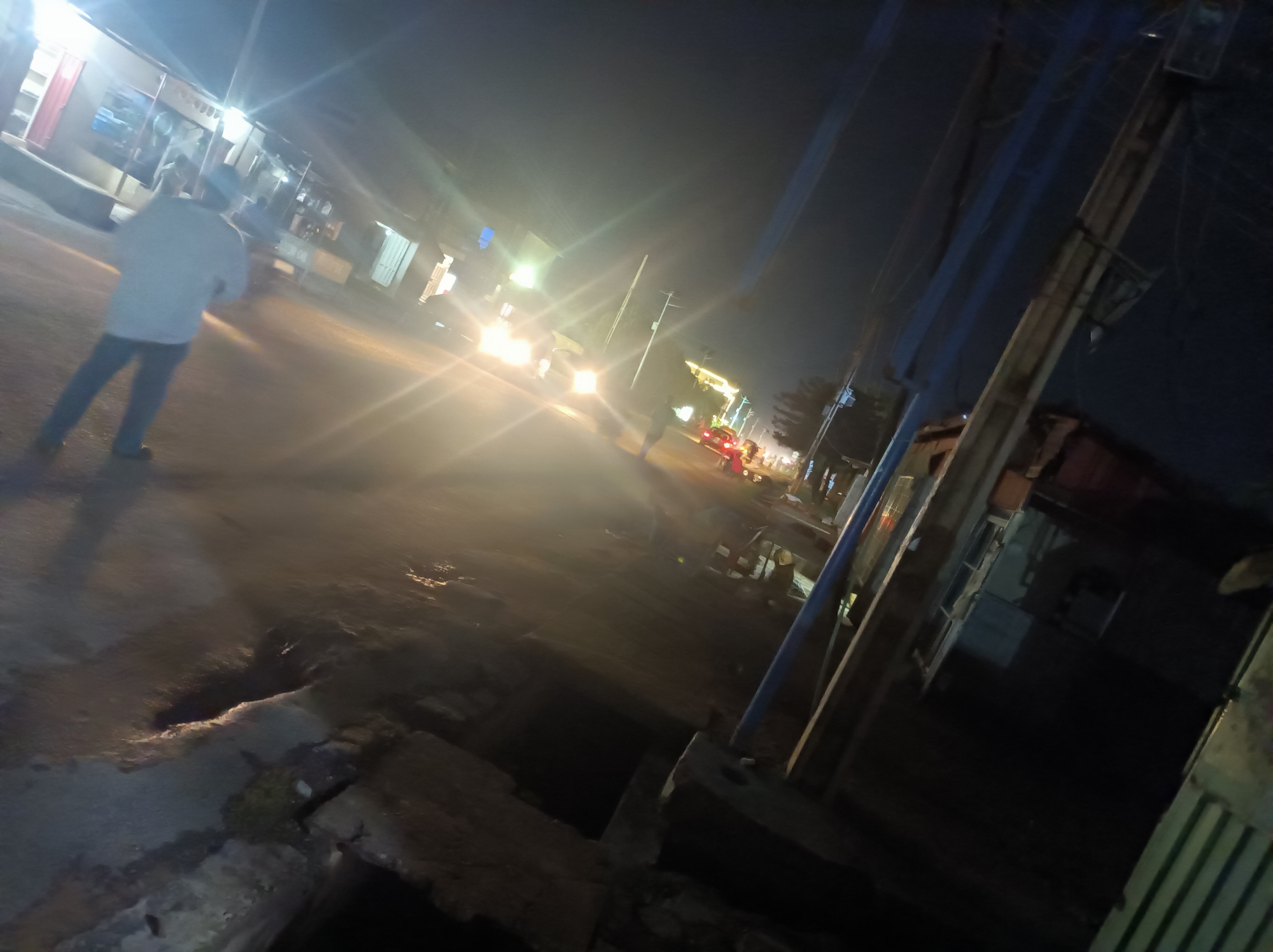 The image shows a dimly lit street scene at night in Narayi, Nigeria. A man in a light-colored shirt and dark pants is walking on the left side of the road. Several cars with headlights on are moving further down the street. Buildings line both sides of the street. Power lines and utility poles are visible on the right side of the frame. The foreground shows a rough, broken pavement. The overall light is low, with glare from the car headlights.