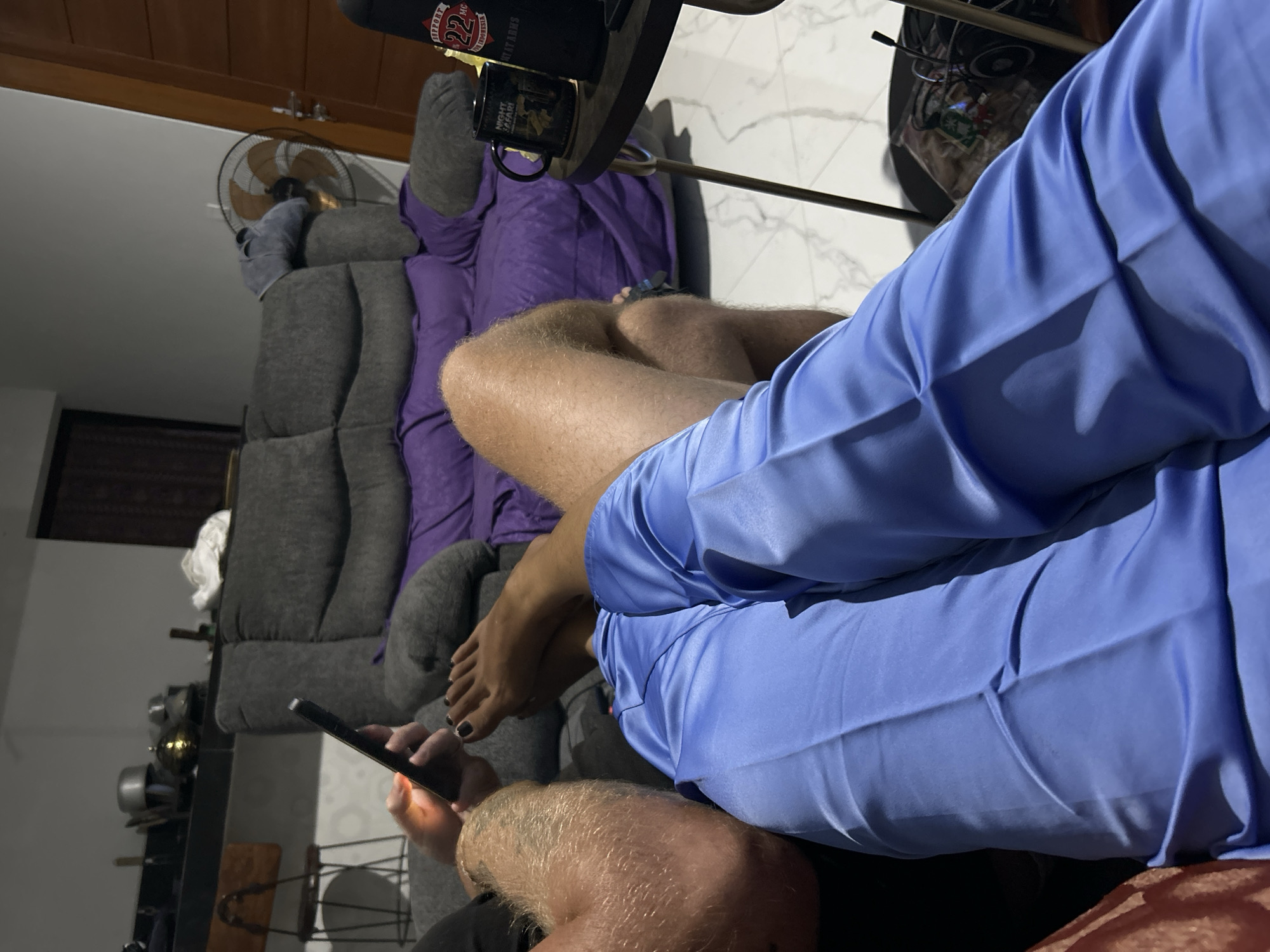 The media file shows a close-up view of people and objects in a room, likely in Sukawati, Indonesia. The scene is dimly lit, suggesting it might be evening or nighttime.
In the foreground, a person's legs are visible, covered by a light blue collared shirt. Their left leg is bent, and their bare foot, with dark nail polish, is resting on a gray couch. Another person's hairy legs are also visible in the lower part of the frame, suggesting two people are present.
A dark-colored smartphone is being held by a hand with dark nail polish, illuminated by its screen, indicating someone is actively using it. The phone is positioned between the two individuals.
The background features a plush gray sofa draped with a purple blanket. A black mug with an image and text is placed on a round table, along with other objects that are not clearly discernible. A ceiling fan is mounted on the wall. The floor appears to be tiled with a white and gray marble pattern.
On the table, a black object with a red and white logo is visible, with text that reads "SUPPORT 22MG" and "INDONESIA". The mug on the table has the text "NIGHT SAFARI" visible on it. The overall atmosphere appears relaxed and casual, with the individuals seemingly engaged in personal activities.