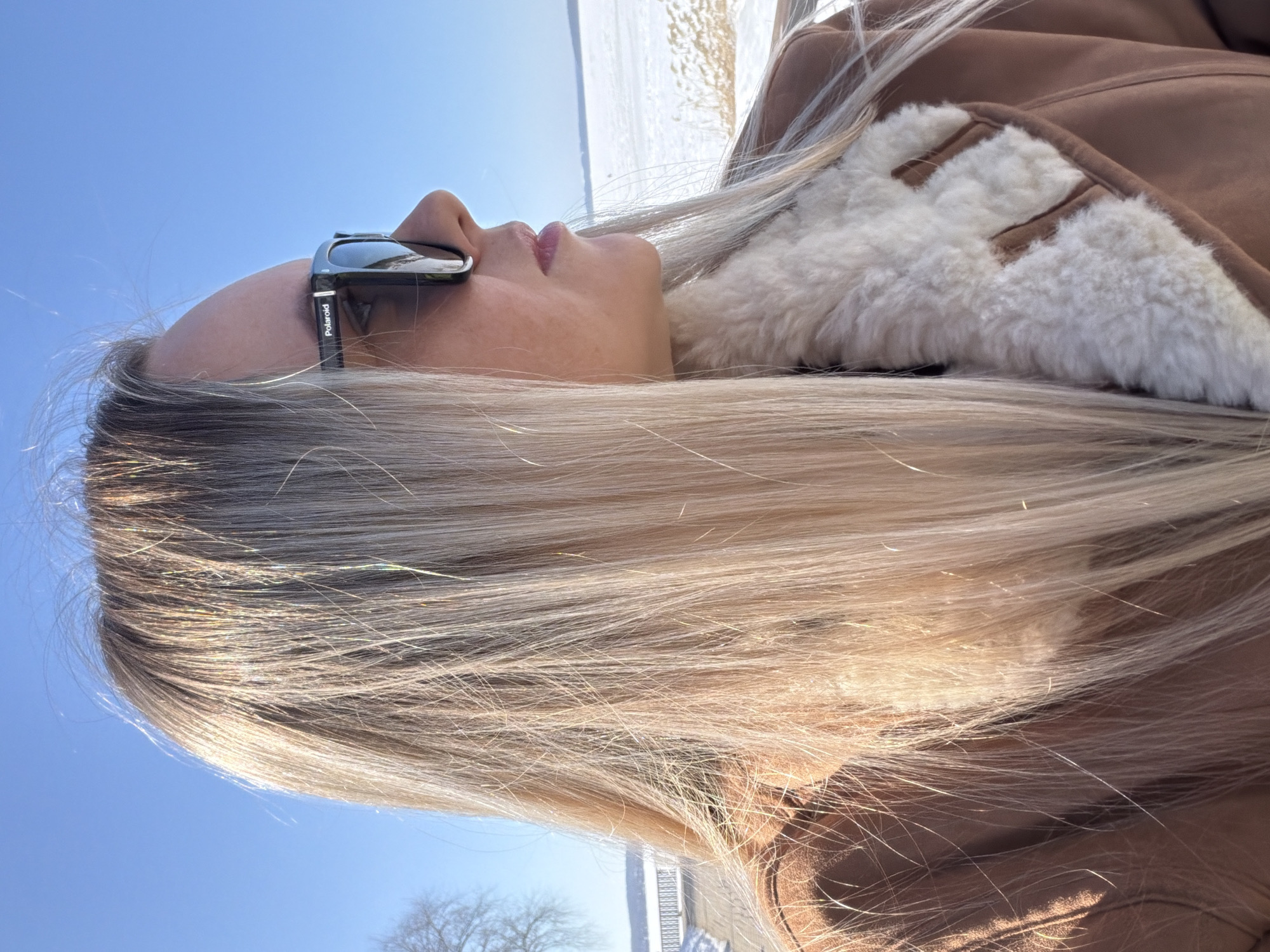 A female subject is depicted from the mid-chest up in a profile view, facing towards the right against a bright outdoor winter landscape in Votkinsk, Russia. She has long, straight, blonde hair, illuminated by strong sunlight which creates prominent highlights. She is wearing black "Polaroid" brand sunglasses that obscure her eyes. Her mouth is closed, with subtly tinted lips. She is dressed in a brown, possibly shearling-lined, jacket or coat, with a cream-colored, textured faux sheepskin or shearling collar and interior visible.
The background consists of a clear, bright blue sky in the upper portion. To the left, a bare-branched tree is partially visible. The mid-ground features a dark-colored pathway or promenade, with a low white barrier or railing along its edge. Beyond the pathway, a vast, flat expanse of snow-covered terrain, likely a frozen body of water such as the Votkinsk pond or reservoir, stretches into the distance under the clear sky. Patches of light brown, dry vegetation are visible along the snow's edge in the lower right. The scene is illuminated by strong natural daylight.