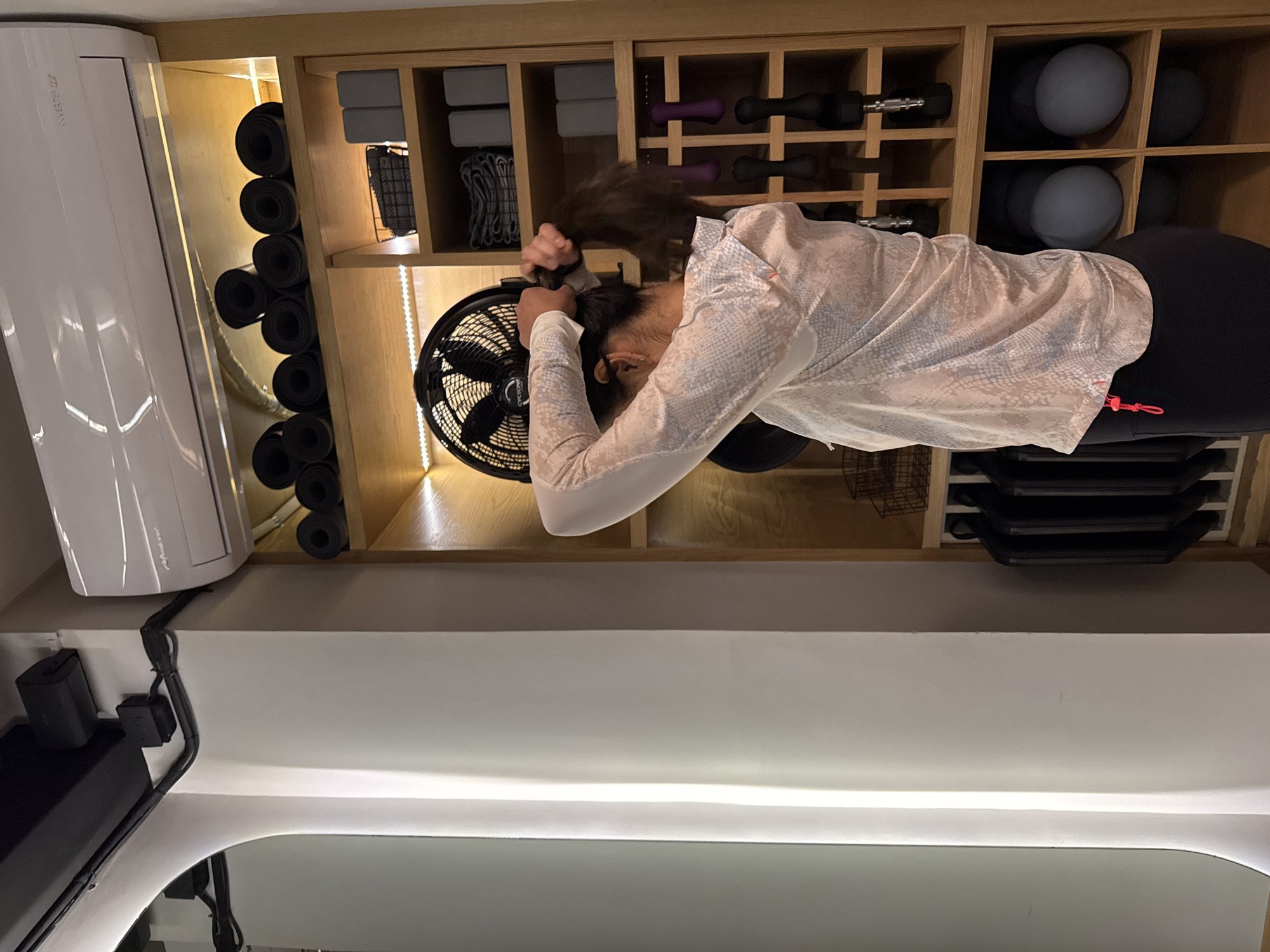 A waist-up view depicts a person in active wear, facing a rotating floor fan with hands raised to tie back their hair. The subject is wearing a long-sleeved, snake-print patterned shirt and black leggings, positioned in front of a shelving unit stocked with exercise equipment like yoga mats, blocks, weights, and exercise balls. The setting appears to be a home gym or fitness studio. Additional elements include an air conditioning unit mounted on the wall and overhead lighting. There is no clear evidence indicating the location being Puebla, Mexico.