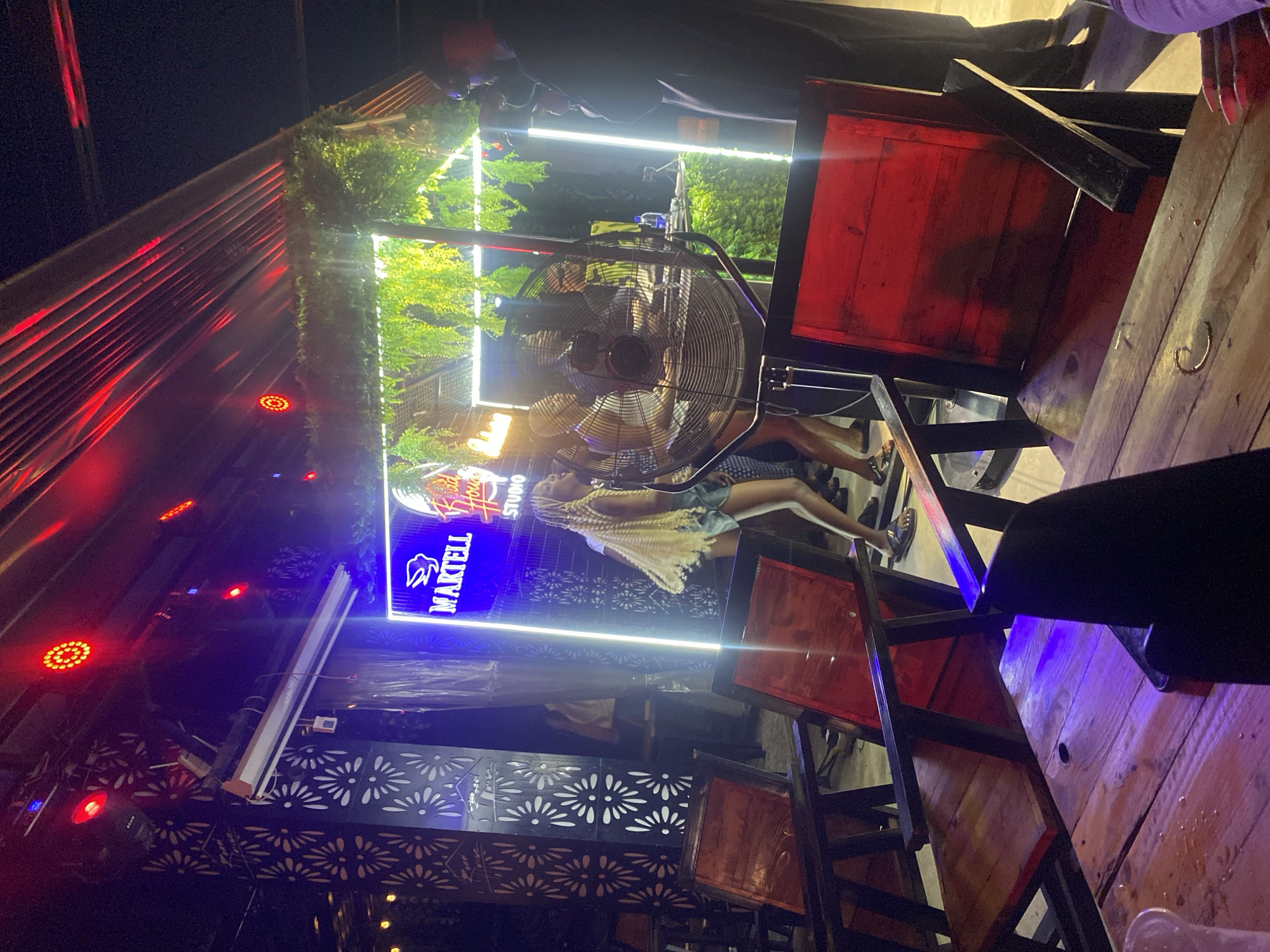 The image depicts an outdoor nighttime scene at what appears to be a bar or club in Howayi, Nigeria. The environment is lit by various artificial lights, including red and blue neon signs, and bright white LED strip lights. In the background, a neon sign displays "MARTELL Food House STUDIO." There is decorative greenery hanging above the illuminated signage. In the mid-ground, a large industrial fan is positioned prominently, with people dancing or standing behind it. To the left, decorative metal screens with floral patterns are visible on a column. The foreground features wooden tables and chairs, with some of the chairs having red wooden seats and black frames. A person's hand with long fingernails is visible in the lower right corner, suggesting they are seated at one of the tables. The overall atmosphere is lively, characteristic of a nighttime entertainment venue.