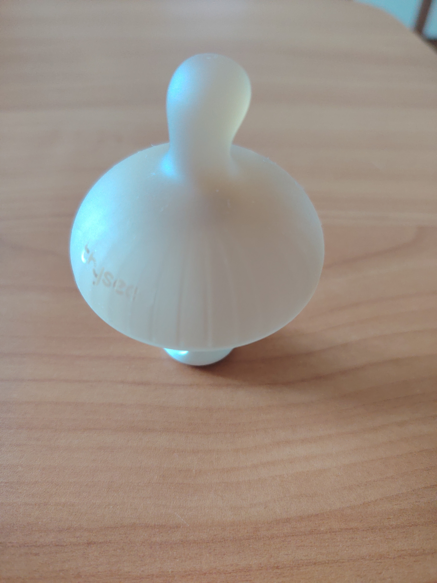 A white, matte-finished object with a rounded body and a narrow neck is centered on a light brown, wood-grain surface. The object is illuminated from the left, casting a soft blue-green sheen on its surface. The word "physiod" is faintly visible etched into the object's body. The background is out of focus, revealing a textured, possibly wooden, surface. This image was taken in Konakovo, Russia.