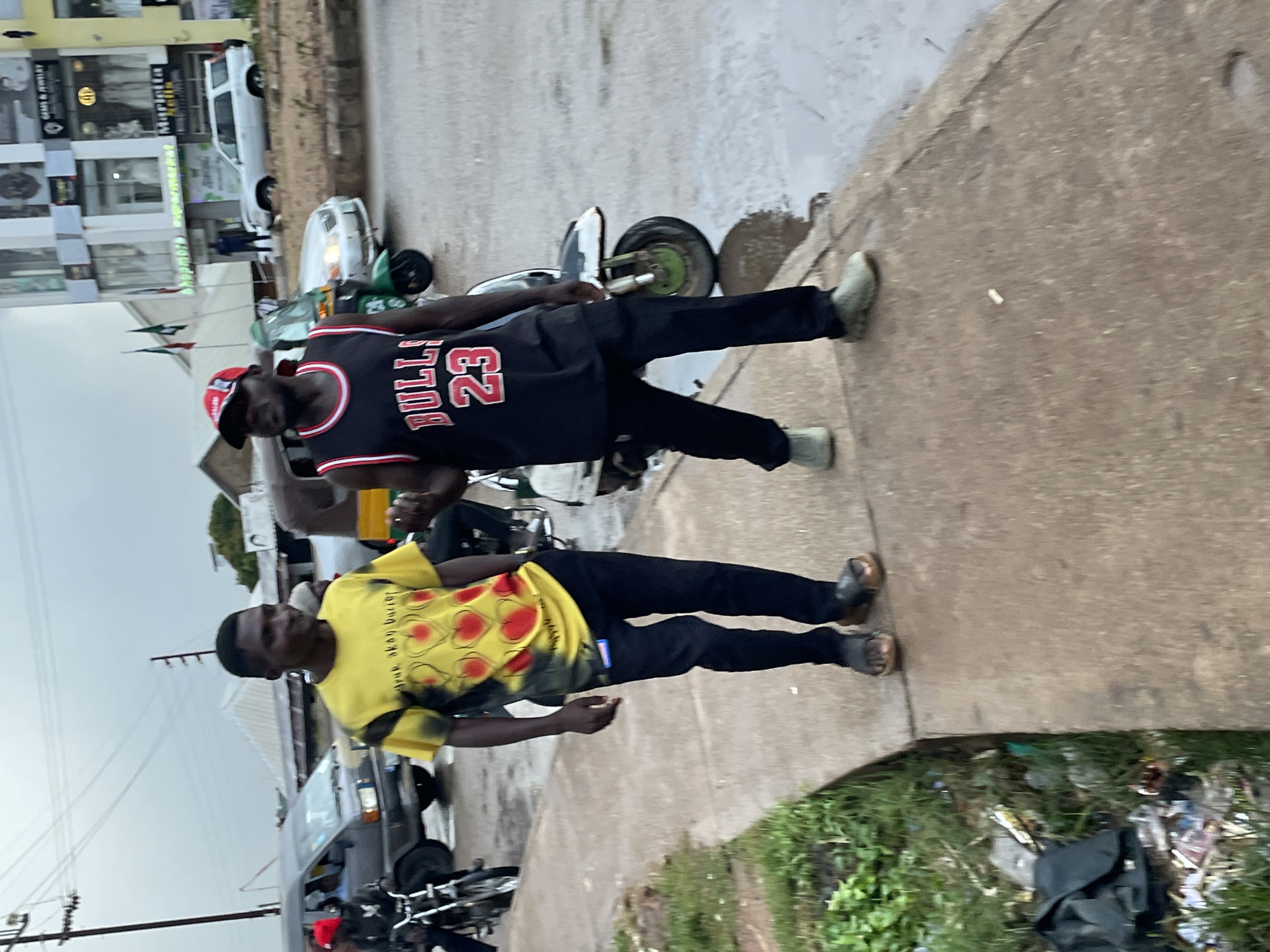 Two men are walking on a sidewalk in the city of Jos, Nigeria. The man on the left is wearing a yellow t-shirt with red heart designs and black pants. He is also wearing sandals. The man on the right is wearing a black Bulls basketball jersey with the number 23, black pants, and light-colored sneakers. He is wearing a red and black baseball cap. They are walking towards the viewer. The sidewalk is made of concrete and is wet, indicating it may have rained. There is a grassy area with litter to the left of the sidewalk. In the background, there are vehicles, including a van and a tricycle, as well as buildings. The sky is overcast.