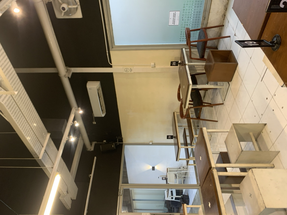 The image shows the interior of a cafe in Malang, Indonesia, located at latitude -7.941 and longitude 112.608. There are several tables with white metal frames and wooden tops, arranged in a square formation. The tables are surrounded by white metal chairs with wooden seats, except for one with a dark brown chair. The cafe features a white ceiling with exposed white pipes and a white air conditioner mounted on a black wall. The cafe is empty, with no people present. The walls are painted in yellow and black tones. Two windows are visible. The window on the left side looks into another room with a couch and another table with chairs. The window on the right side is frosted. The table in the foreground has a sign with the number "19" on it. There is a sign on the wall beside the window on the right with an inscription in Indonesian. The floor is tiled white.