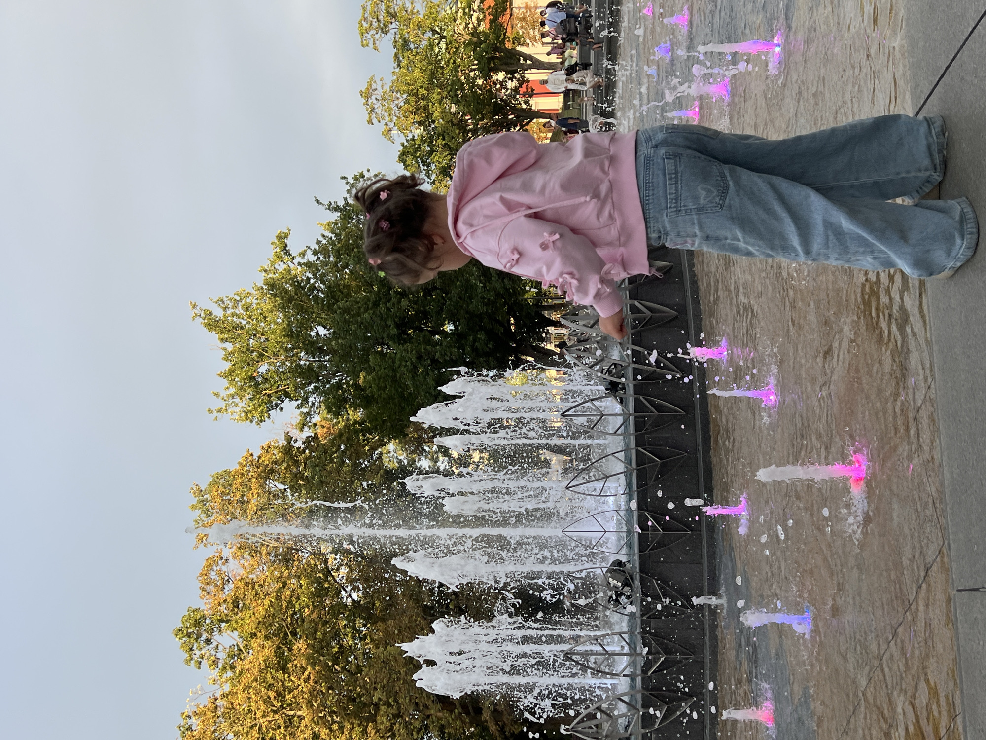 A young girl in a pink hooded jacket and wide-leg jeans stands facing away from the camera, playing with a fountain in a public square. The fountain features several large jets of water in the background and smaller, colored jets in the foreground, illuminated with pink and blue lights. Trees with yellowing and green leaves line the background, suggesting it is autumn. In the far background, other people are visible in the plaza, some seated on benches. The ground is paved. The image appears to be taken in the city of Lublin, Poland.