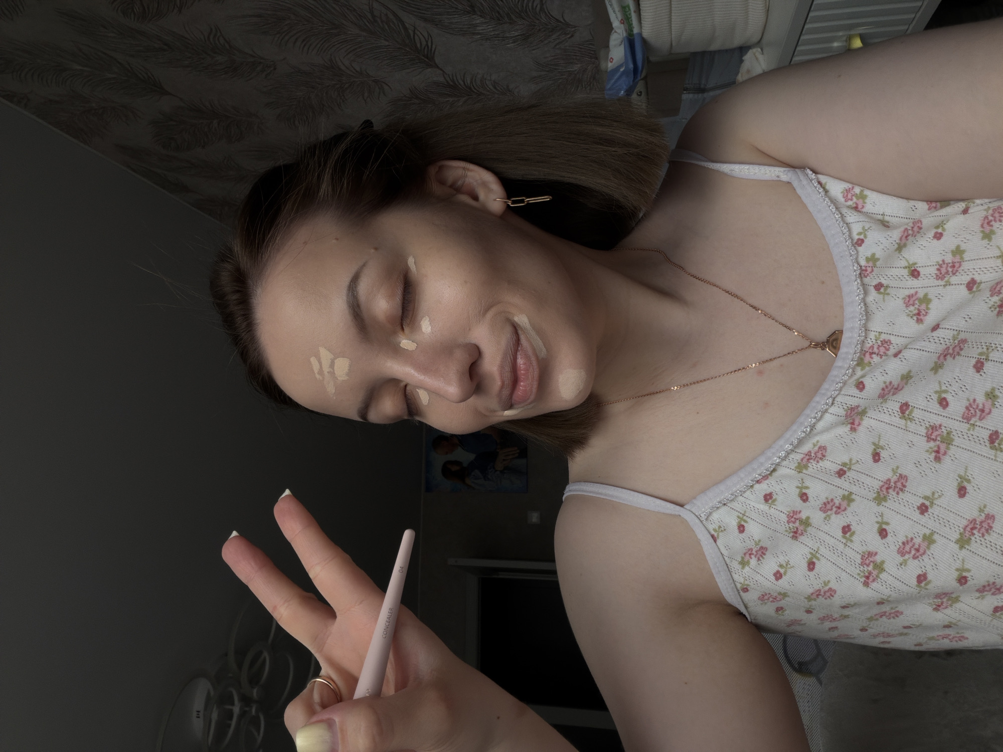 The image depicts a young woman indoors, likely in a private residence, as indicated by the personal items and décor in the background. The woman is facing away from the camera, at an angle to the side, with her eyes closed. She has makeup applied on her face in several spots, and she is holding a makeup brush in her hand, gesturing with a peace sign. She is wearing a floral-patterned tank top. A gold necklace is visible on her neck, and a pair of gold earrings is visible in her ear. The background is a mix of dark and light areas, and a blurry picture can be seen. The lighting is somewhat dim. There is no definitive visual indication that the photo was taken in Reshetnikovo, Russia.