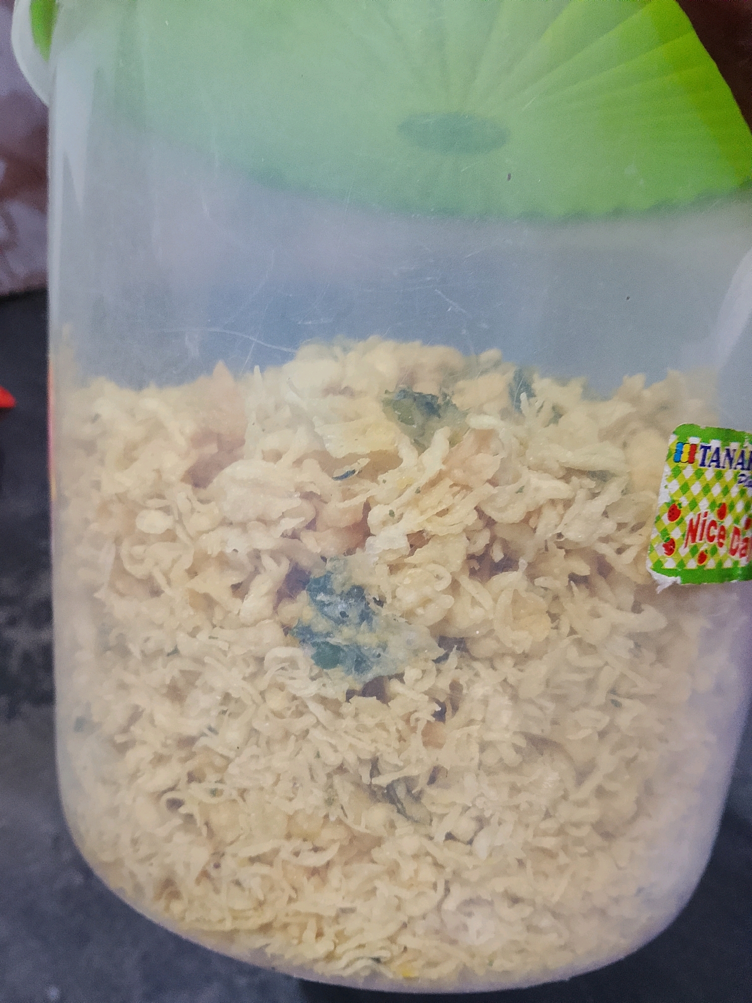 In Labuhan Ratu Sembilan, Indonesia, a clear plastic container with a green lid holds a light-colored, dry food item. The food appears to be composed of small, irregular pieces. Some of these pieces have a slightly darker, greenish hue, suggesting the presence of herbs or spices. A small sticker with green and white checkered pattern and red fruit designs is affixed to the right side of the container, with the text "Nice Day" visible. The container is partially filled with the food item, and the background is blurred, suggesting a shallow depth of field.