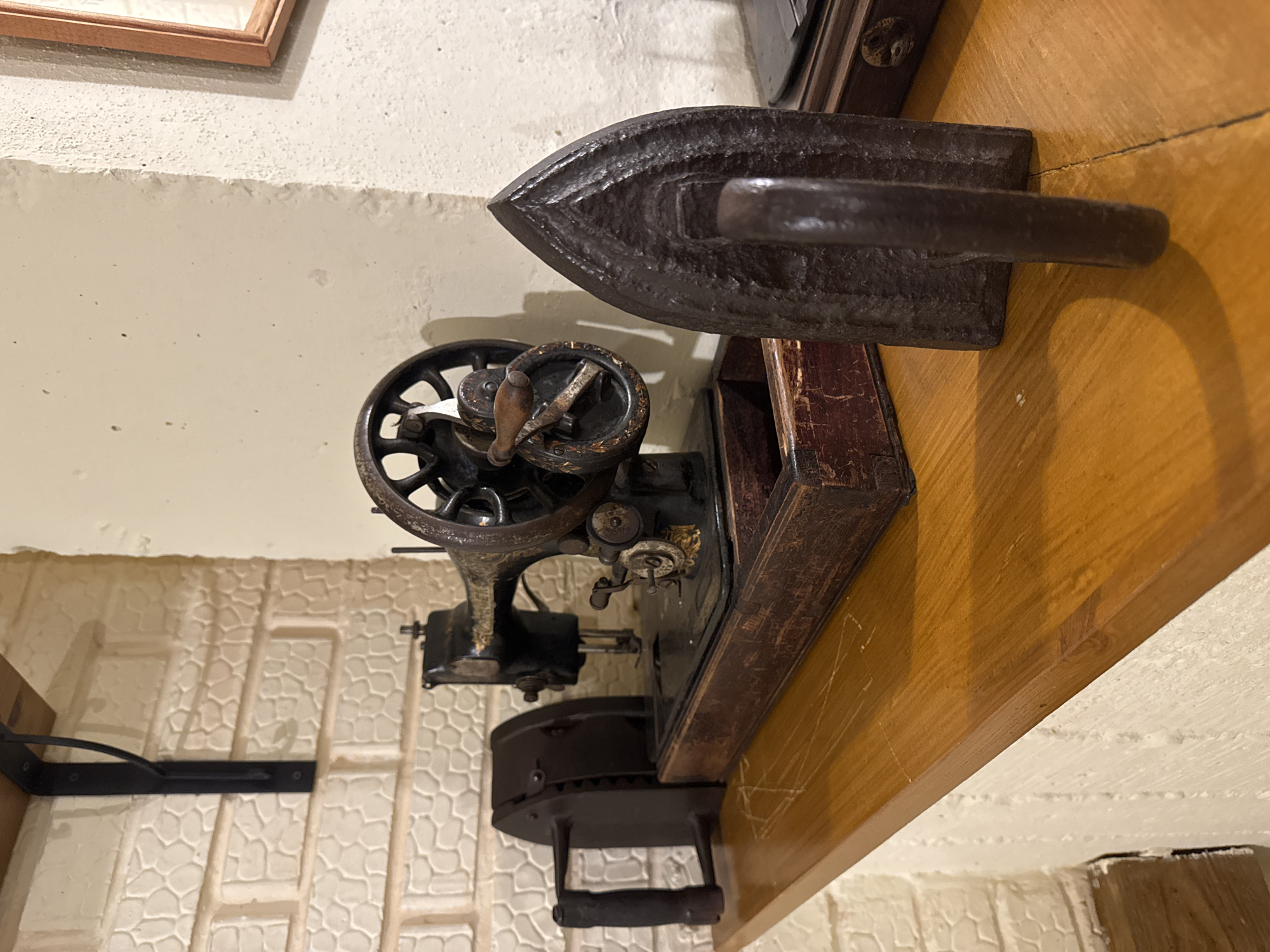 The scene depicts a static display of antique domestic items on a polished wooden shelf within an interior setting in Chaikovsky, Russia. From left to right, the objects are: a dark, cast-iron sad iron with a prominent wooden handle, positioned horizontally on the shelf; an antique, black metal sewing machine, likely cast iron, with a large handwheel, golden decorative elements, and mounted on a dark, distressed wooden base with a visible front compartment; and a second dark, cast-iron sad iron, positioned vertically with its pointed end facing the viewer. The background on the left features a light-colored, textured wall resembling irregular brickwork, with a black metal shelf bracket visible above the objects. The background on the right consists of a smoother, light-colored wall, with a partial view of a wooden-framed item in the upper right quadrant. The wooden shelf is reddish-brown, exhibiting a semi-gloss finish and minor surface scratches.