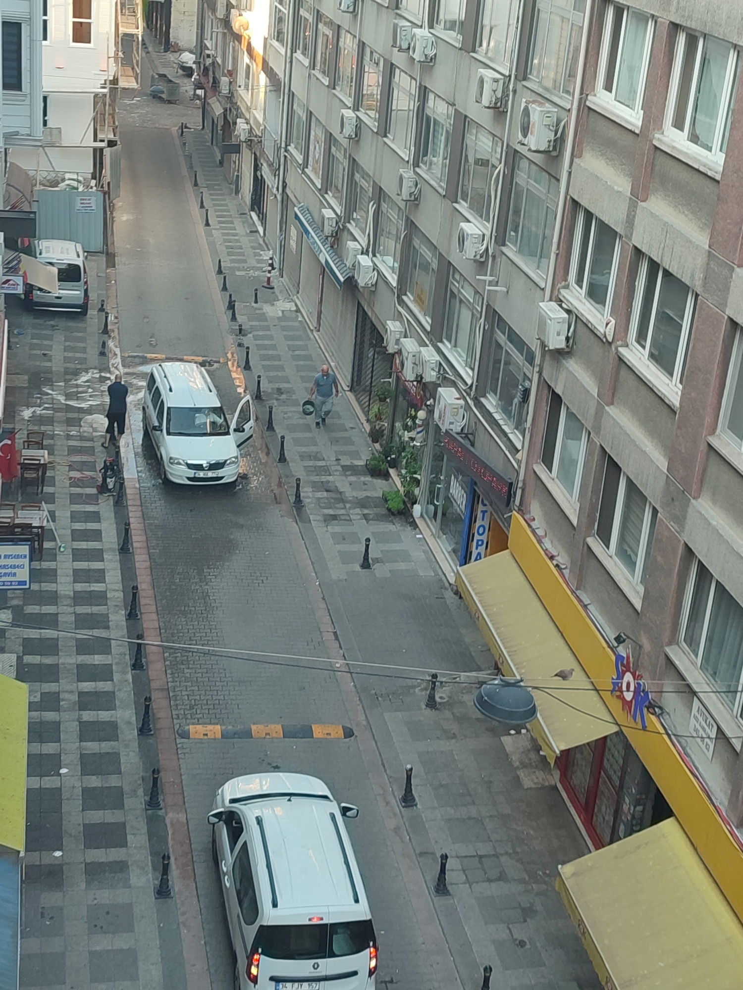The image depicts a street scene in Kadıköy, Turkey, likely during daylight hours, with a narrow street lined by buildings on both sides. The street is paved with cobblestones and has a checkered pattern on the sidewalks.
On the left side of the street, a white van is parked with its rear door open. Two men are near this van. One man, wearing dark shorts and a dark t-shirt, stands with his back to the camera, facing the van. The other man, wearing lighter clothing, is further down the street, walking away from the van. Across from the van, there is an area with tables and chairs, suggesting an outdoor seating area for a business, possibly a cafe or restaurant, with a Turkish flag visible.
On the right side of the street, a multi-story building dominates the view. The building has numerous windows and several air conditioning units mounted on its exterior. There are shops at the ground level, some with awnings. One shop has a sign that appears to say "BARBER" and another with "TOPAZ". Further down the street on the right, another white van is visible, parked in front of a yellow awning. A speed bump is present on the street. A bird is perched on the yellow awning.
The overall impression is an urban street with commercial activity and residential units above. The presence of multiple parked vehicles and pedestrians indicates a moderately busy environment.