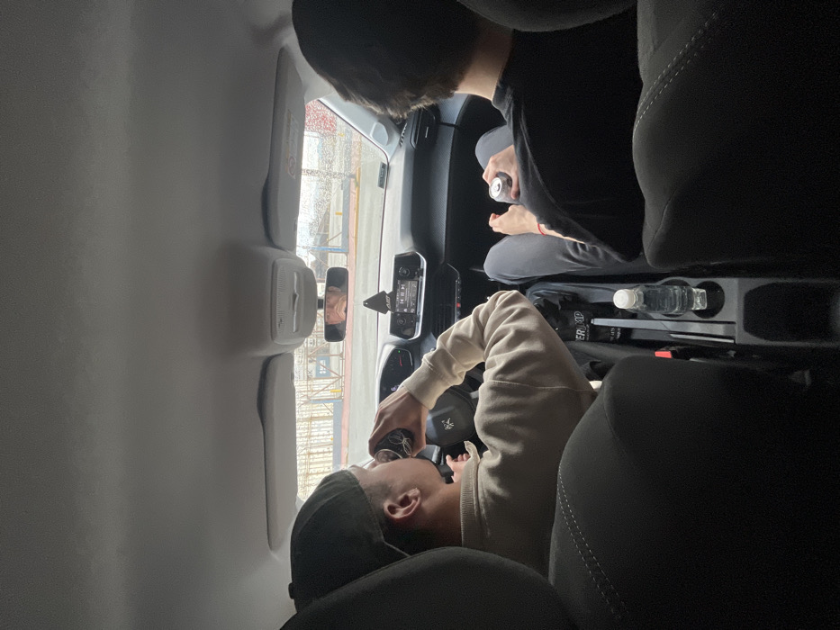 An interior shot from the rear seat of a vehicle captures two individuals in the front seats in Ushuaia, Argentina. The driver, a male wearing a dark baseball cap backward and a light-colored long-sleeved shirt, is actively drinking from a metallic can while his hands are near the steering wheel displaying a Peugeot logo. To his right, the front passenger, a male with dark hair and wearing a dark t-shirt and dark trousers, holds a similar metallic can in his left hand, resting on his lap. A clear plastic water bottle is positioned in a cup holder on the central console between the seats. Through the windshield, an exterior scene depicts a multi-colored metal barrier or fence structure (light blue/white and yellow) suggesting a port or industrial area, with visible water droplets on the glass indicating precipitation. The environment outside appears overcast.