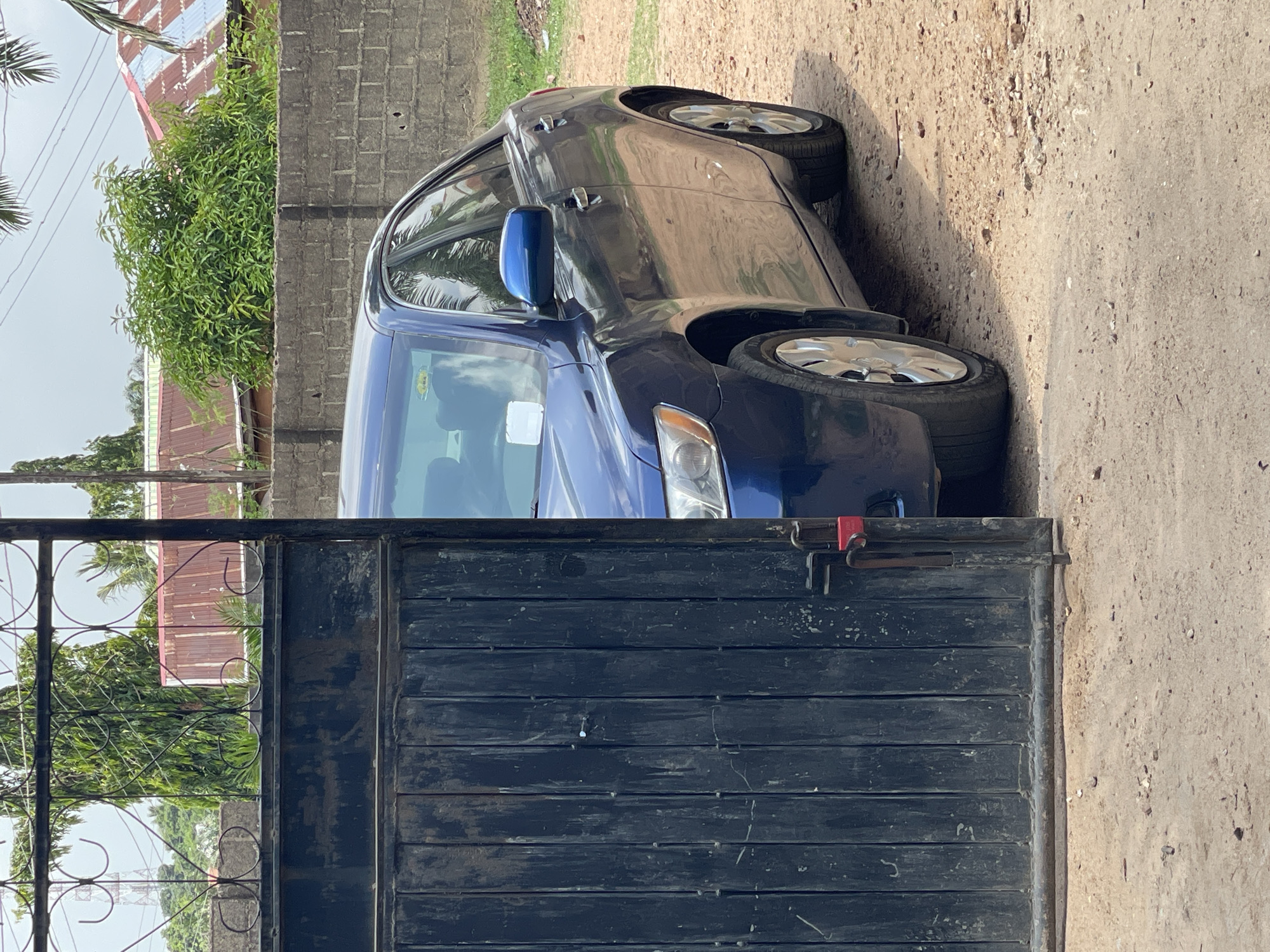 The image shows a blue and gray car parked behind a closed, dark metal gate in Akwanga, Nigeria. The vehicle's front end is blue, while the rear is gray. The gate is constructed of horizontally arranged metal slats. The ground is a mix of dirt and concrete. In the background, there's a brick wall, greenery, and a building with a red and white roof. The photograph appears to have been taken during daylight hours.