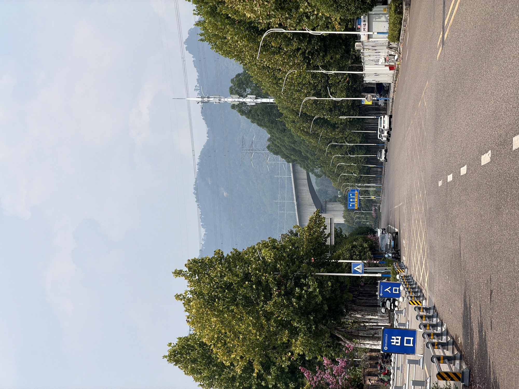 The image depicts a street scene in Jiangjin, China, featuring a road lined with trees. In the foreground, the asphalt road is marked with white dashed lines and yellow double lines. On the left side of the road, there are metal barriers with yellow and black striped poles, and a blue sign with white Chinese characters that reads "出口" (Exit). Further along the left, there are parked motorcycles and a dark-colored SUV. A pedestrian crossing sign is also visible. On the right side of the road, a row of streetlights with curved white poles lines the way, and a white structure with corrugated metal siding is present. In the background, a large bridge arches over the road, and a cellular tower stands tall amidst a backdrop of lush green hills and a hazy sky. The overall atmosphere suggests a relatively quiet urban environment with natural elements incorporated into the infrastructure.