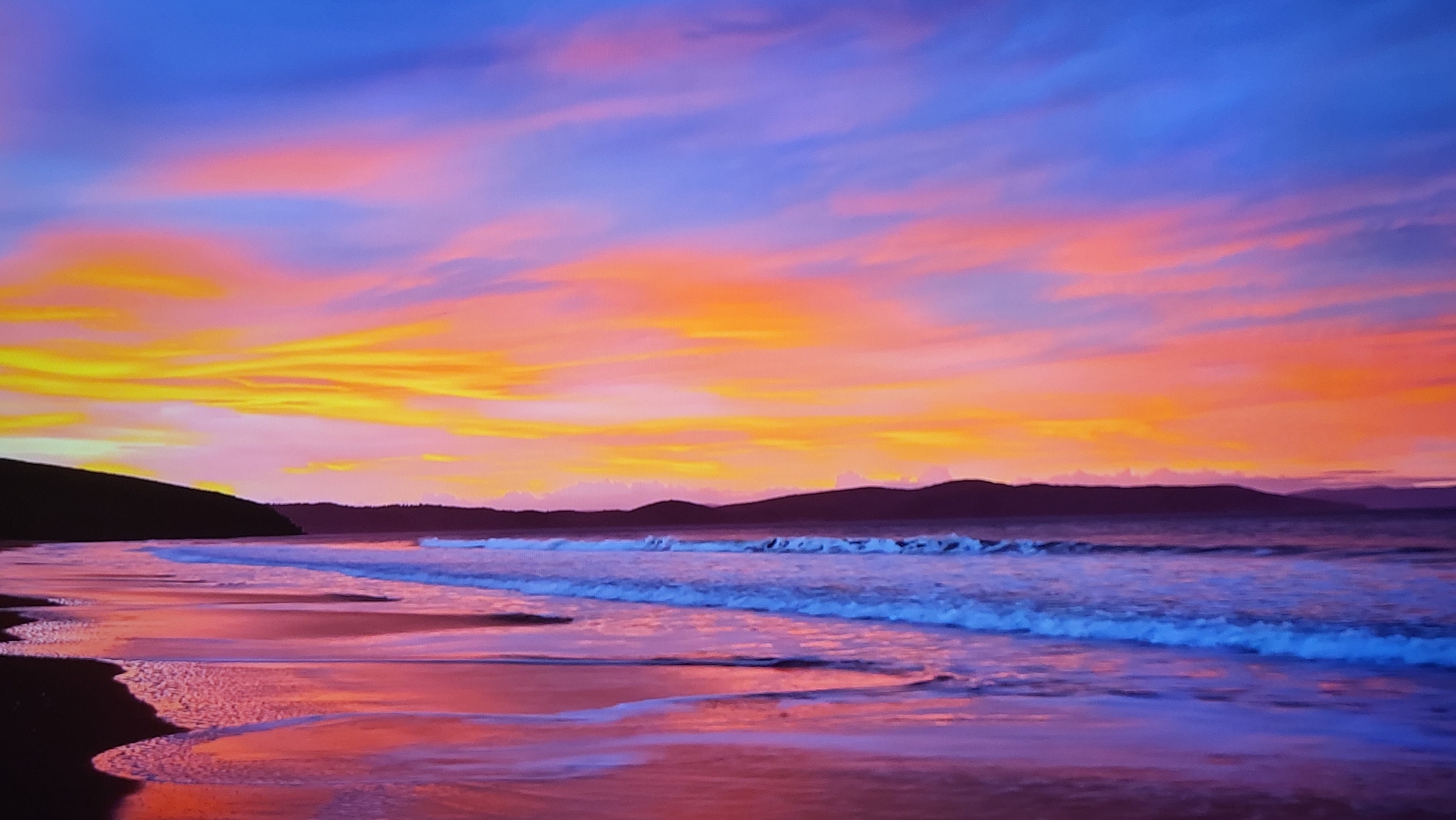 The image depicts a scenic coastal landscape at sunset. The sky is a vibrant mix of colors, transitioning from deep blue at the top to shades of pink, orange, and yellow closer to the horizon. The sun's light illuminates the clouds, creating a dynamic and colorful display.
Below the sky, there is a body of water, likely the ocean, with gentle waves approaching the shore. The waves have white foam crests. The beach is visible in the foreground, with the wet sand reflecting the colors of the sky, creating a mirrored effect.
In the distance, there are dark, silhouetted landmasses or mountains, providing a backdrop to the scene. The overall impression is one of tranquility and natural beauty.
The image does not appear to be in Yerevan, Armenia, which is a landlocked city without a coastline.