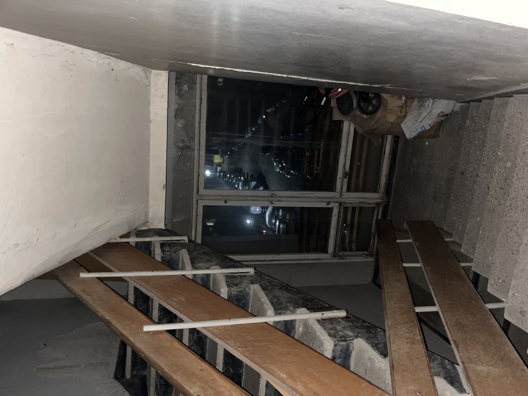 The image captures an indoor scene, likely a stairwell or landing in an older building in Sofia, Bulgaria, characterized by a utilitarian and somewhat unfinished appearance. The perspective is tilted, showing the scene as if viewed from an unusual angle.
In the center of the frame is a large, multi-pane window, appearing to be made of wood, through which a dark, nocturnal exterior is visible. Distant artificial lights from outside reflect on the window panes. On a ledge or platform to the right of the window, various objects are stored, including part of a stroller or dolly with visible wheels, cardboard boxes, and a crumpled white plastic bag. The bag has some faint, illegible text or a logo.
To the left of the window, a light-colored wall with an uneven, possibly plastered or concrete surface, shows signs of cracking and wear. Above, the ceiling is similarly unadorned concrete or plaster.
The right side of the image features a concrete staircase, with individual steps visible. Laid across these steps are long, rustic wooden planks, secured by horizontal white metal bars. This arrangement suggests a temporary ramp for moving items or protection for the stairs, rather than a permanent fixture. The overall lighting is dim, indicating either night or poor interior illumination. No people are visible, and there is no discernible activity taking place. The scene conveys a sense of a neglected or functional space, possibly awaiting renovation or used for temporary storage.
