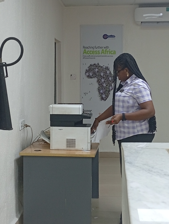 An indoor scene captures a woman engaged in an office task, likely at a service center or bank. The woman, positioned on the right side of the frame, has long, dark braided hair styled over her shoulders. She is wearing a light-colored (possibly light purple or blue) checkered short-sleeved shirt with a collar and dark trousers. She stands facing a white and black multi-function printer/copier, which rests on a desk with a light brown top and a dark grey base. Her hands are actively involved with the machine, with her right hand extended towards the paper tray or output area, and her left hand holding a stack of white papers.
Behind the printer, a beige landline telephone is partially visible, connected to a power outlet on the white wall with visible cords. On the far left, a portion of a black, curved object (possibly a light fixture or speaker) hangs down.
The background features a large poster mounted on the white wall. The poster displays a stylized key logo at the top and text that reads: "Reaching further with Access Africa." The words "Access Africa" are highlighted in a vibrant green font. Below this, smaller text reads: "Enjoy swift and convenient transfers across Africa and beyond." Below the text, there is a graphic representation of the continent of Africa, formed by numerous small, scattered dots or shapes. High on the wall, above the poster, a white air conditioning unit is visible. To the right of the woman, in the foreground, a corner of a white surface with a marble-like pattern is visible, likely another counter or desk.
The setting appears clean and well-lit, suggesting a professional environment. The floor is a light, neutral color. The overall impression is that of an employee performing a routine administrative task in an establishment that offers financial or transfer services related to Africa.
