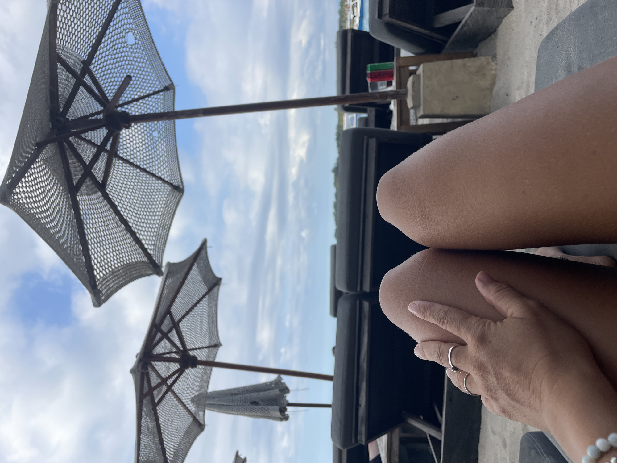 An individual's legs are prominently displayed, resting on a beach lounger. Their hand, adorned with rings, is gently placed on their left thigh, with a beaded bracelet visible on their wrist. In the background, multiple dark beach loungers are arranged on the sand. Overhead, two woven umbrellas cast shadows, and the sky is a mix of blue and scattered clouds. Further in the distance, palm trees and some boats on the water are visible, suggesting a tropical coastal location. The scene appears to be a relaxing day at the beach in an unknown location in Indonesia.