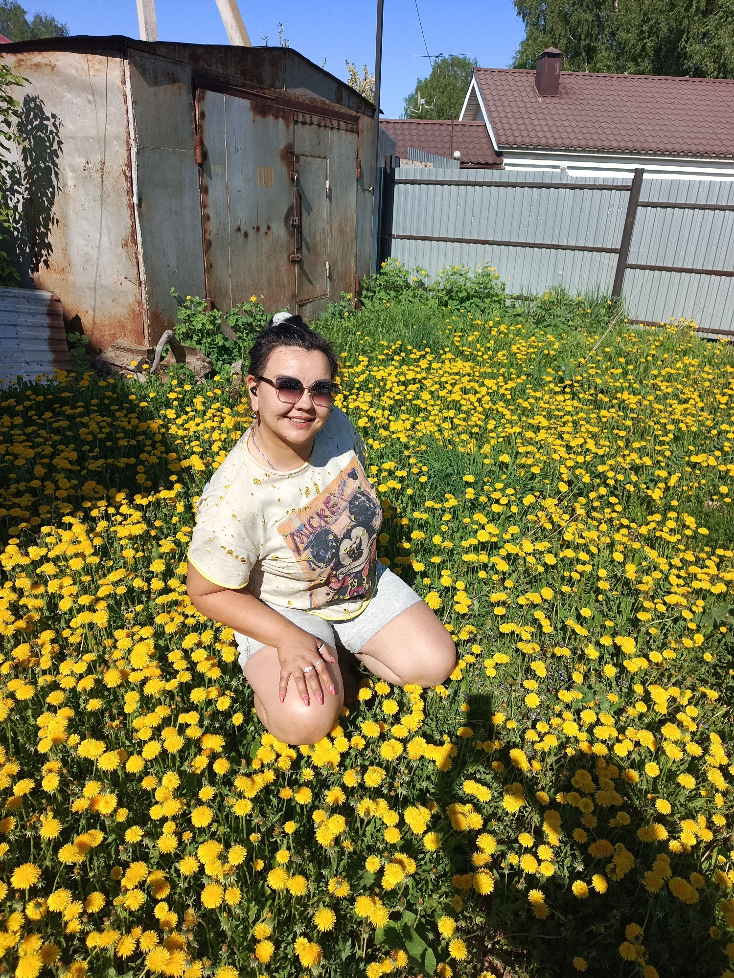 The image shows a woman kneeling in a field of dandelions in Chaikovsky, Russia. She has dark hair pulled into a bun, is wearing sunglasses and a t-shirt with a Mickey Mouse graphic, and gray shorts. A weathered, gray metal garage with rusting seams is in the background, to the left of the woman. Behind the garage, green foliage is visible. To the right of the woman is a gray metal fence and a house with a brown roof. The day appears bright and sunny. The subject is kneeling amidst a dense field of yellow dandelions, with some patches of green grass visible between the flowers. The shadows cast on the field suggest the sun is shining from the right.
