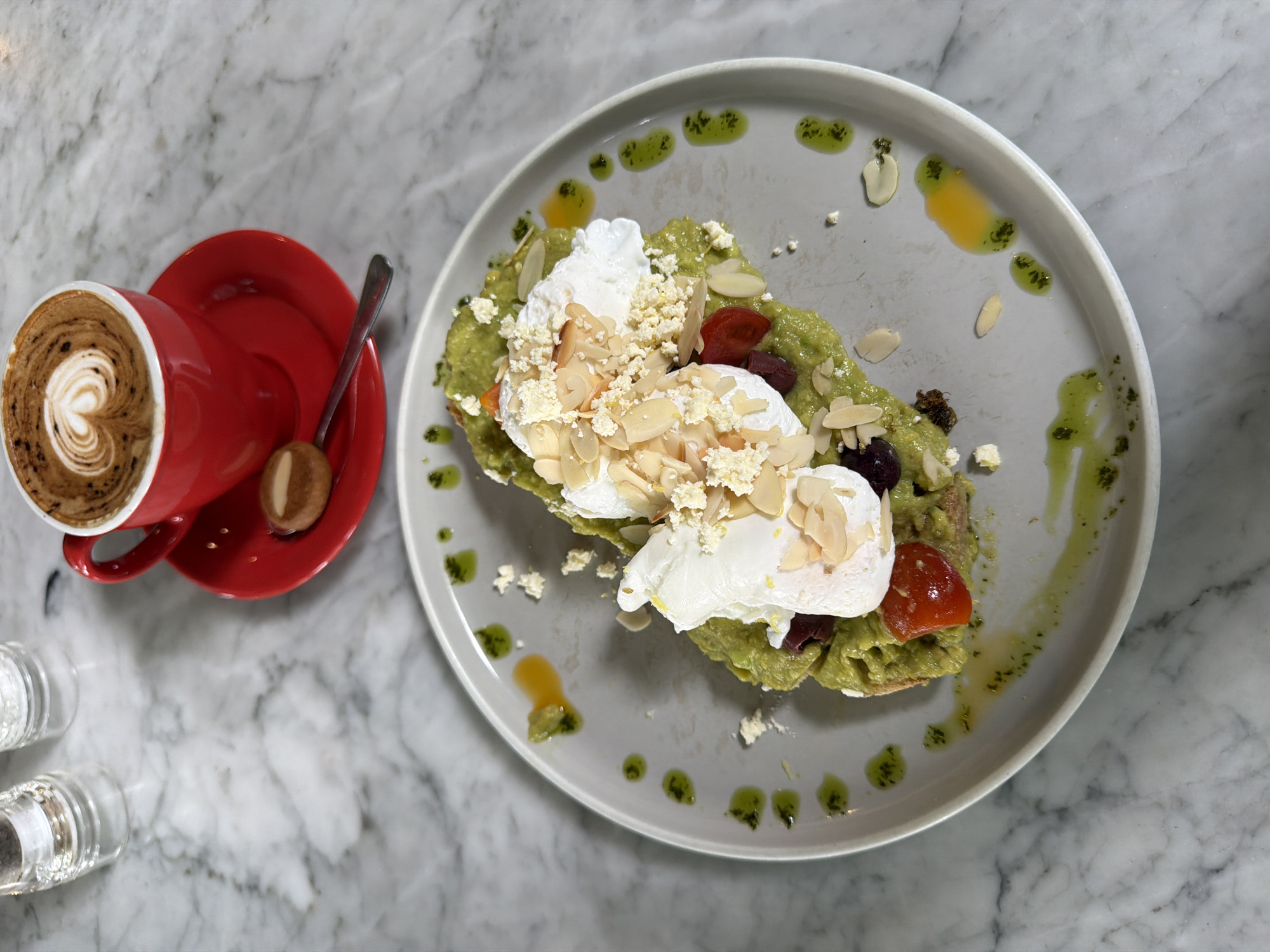 A high-angle shot shows a plate of avocado toast with poached eggs and a cup of coffee, set on a marble countertop. The avocado toast is topped with feta cheese, slivered almonds, and what appear to be cherry tomatoes and black olives. A light green drizzle or sauce decorates the edge of the plate. To the left, a red cup with latte art is placed on a matching red saucer, along with a small, round biscuit and a spoon. In the lower left corner, the tops of two glass containers, potentially salt and pepper shakers, are visible. The marble countertop has a grey and white swirling pattern. The image was likely taken in North Kuta, Indonesia.