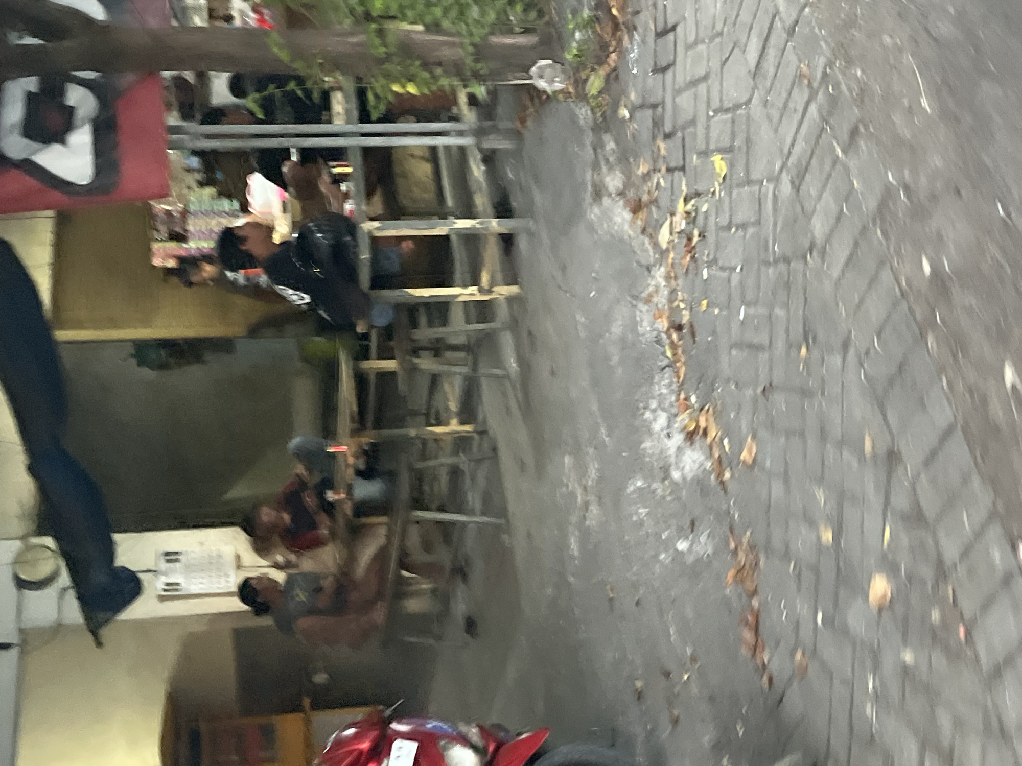 In Gelam, Indonesia, an outdoor seating area at what appears to be a food stall or small restaurant is captured in the image. Several individuals are seated at wooden tables, some engaged in conversation or eating. A red motorcycle is partially visible on the left side of the frame. The ground is paved with interlocking bricks, and scattered dry leaves suggest an outdoor setting. In the background, there are structures with awnings and a sign with graphic elements. The lighting is dim, indicating it may be evening or an overcast day.