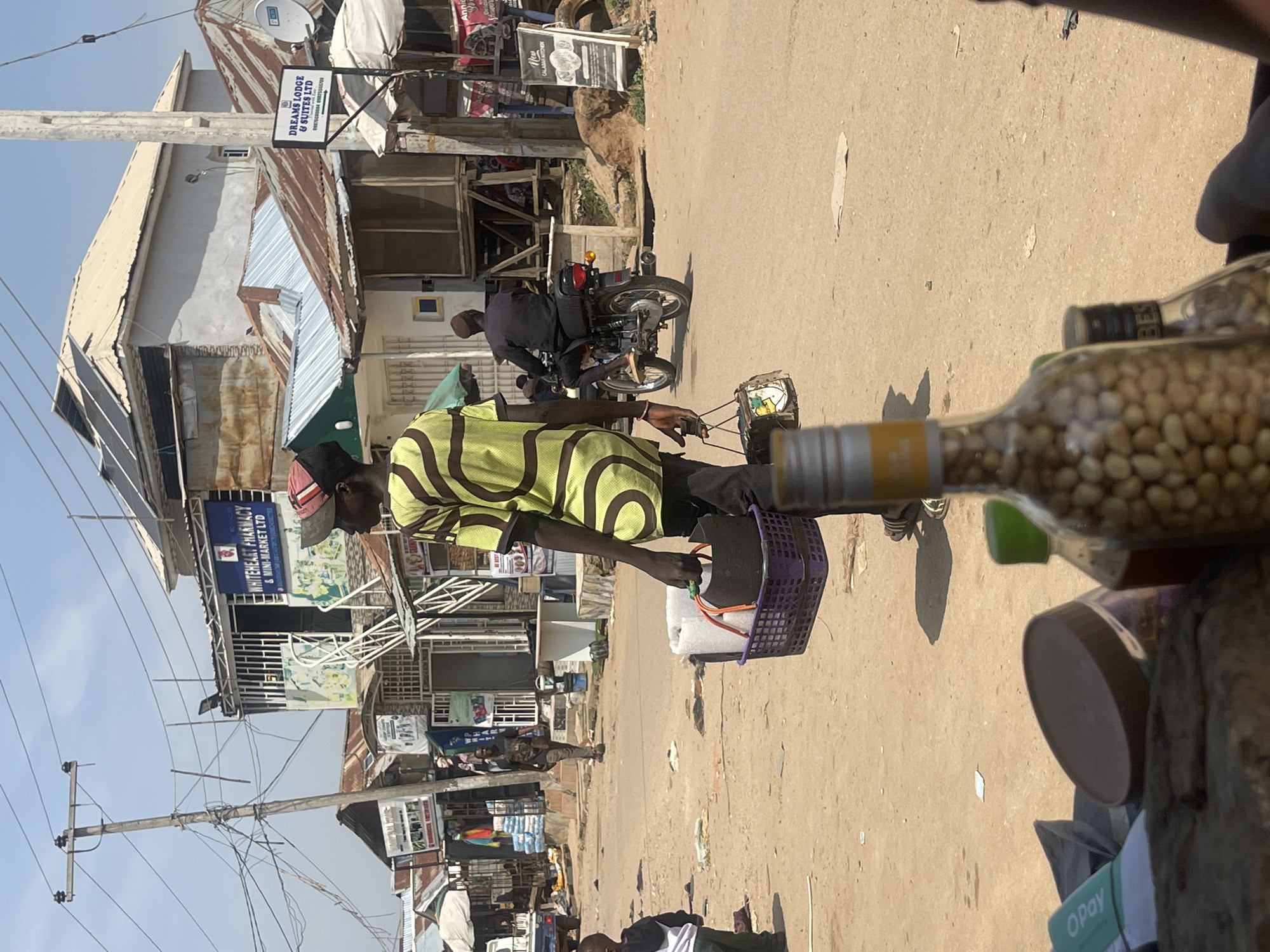 The image depicts a street scene in Abuja, Nigeria, under a clear sky. In the foreground, a person walks away from the camera, carrying a purple basket filled with items, including what appears to be rolls of fabric or padding, and a dark, round object, possibly a pot or bowl. The person is wearing a yellow and black patterned shirt, dark pants, and a red and black cap. They are also carrying a small, patterned bag in their left hand. In the middle ground, a man is riding a motorcycle away from the camera. Behind him, several buildings line the street, some with corrugated iron roofs and others with more modern construction, including one with solar panels. Signs for "WHITEHEART PHARMACY & MINI-MARKET LTD" and "DREAMS LODGE & SUITES LTD" are visible on the buildings. The street itself is unpaved and dusty, with scattered debris. A young child in a white shirt and dark pants stands in the left middle ground, facing away from the camera. Further in the background, other people and vehicles are present, suggesting a bustling marketplace or residential area. The overall impression is a snapshot of everyday life in a Nigerian city.